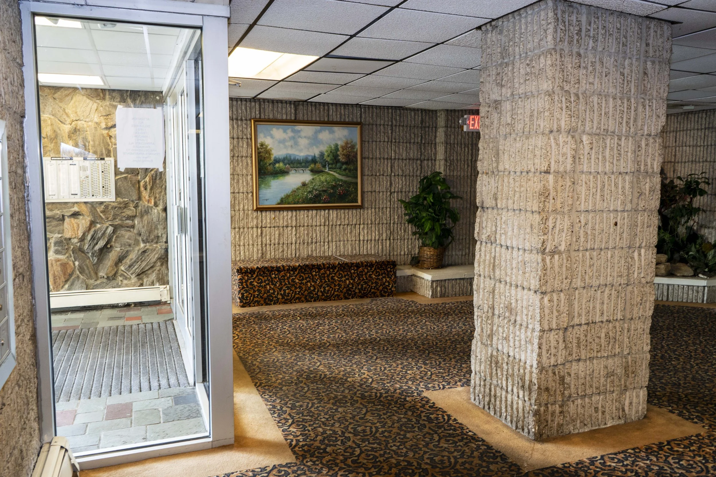 Interior view of a building lobby with a glass door, stone and brick walls, a patterned rug, a framed landscape painting, and potted plants.
