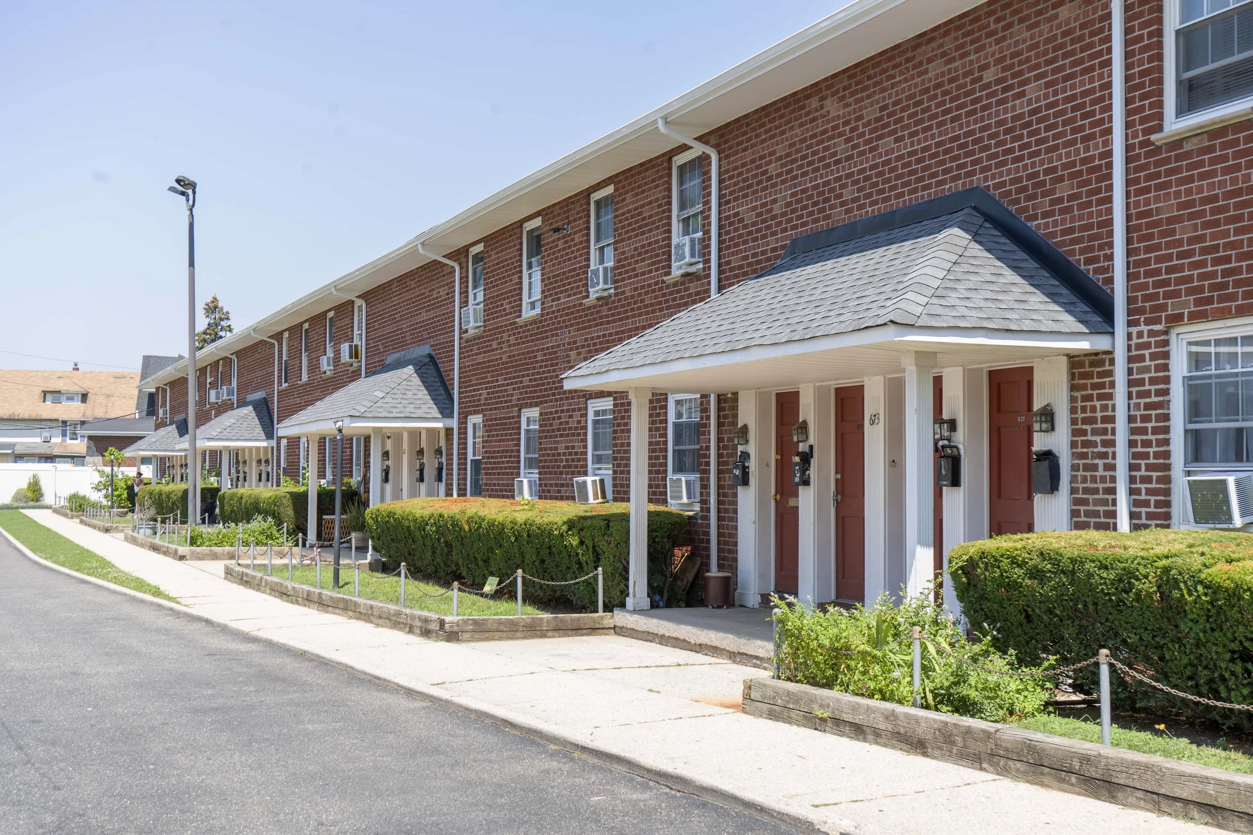 Row of red brick apartment buildings with small front porches, bushes, and walkways.