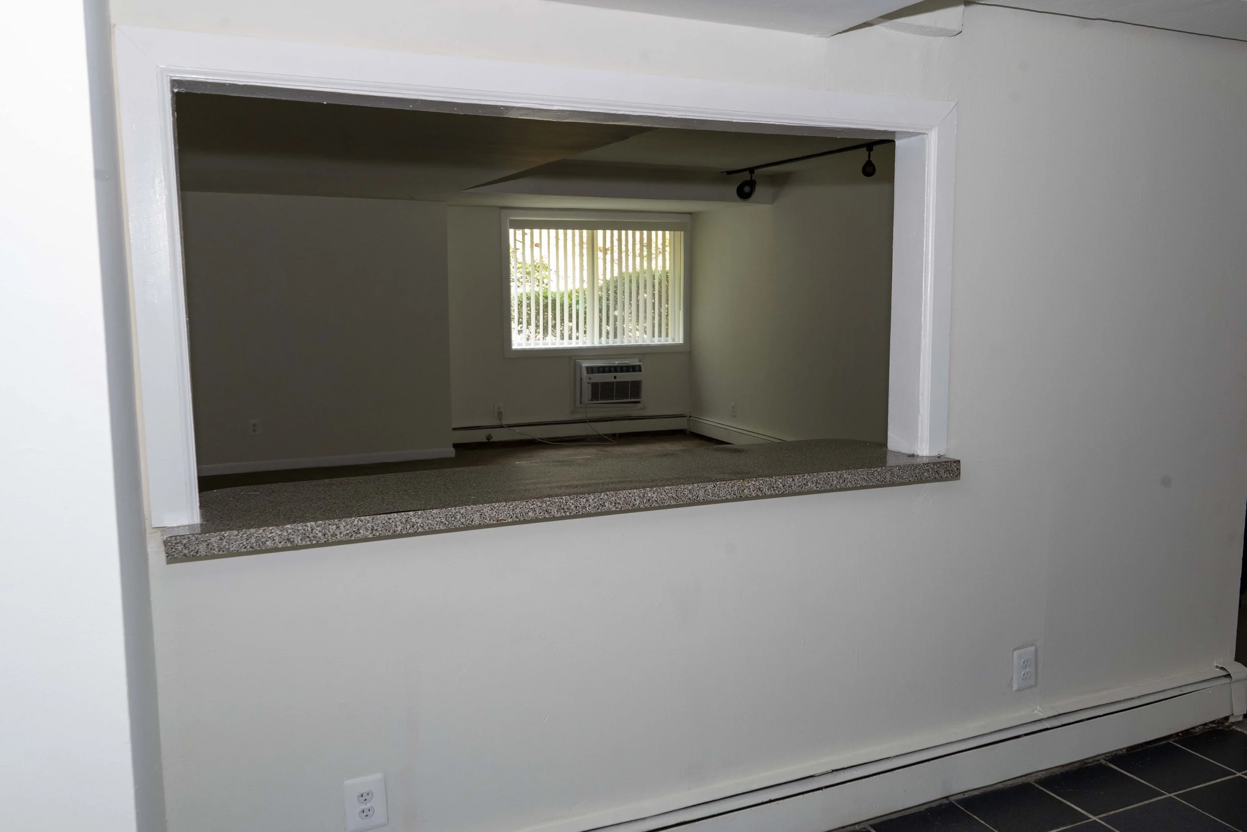View of an empty room seen through a kitchen pass-through with a granite countertop, beige carpet, window with vertical blinds, and an air conditioning unit below the window.