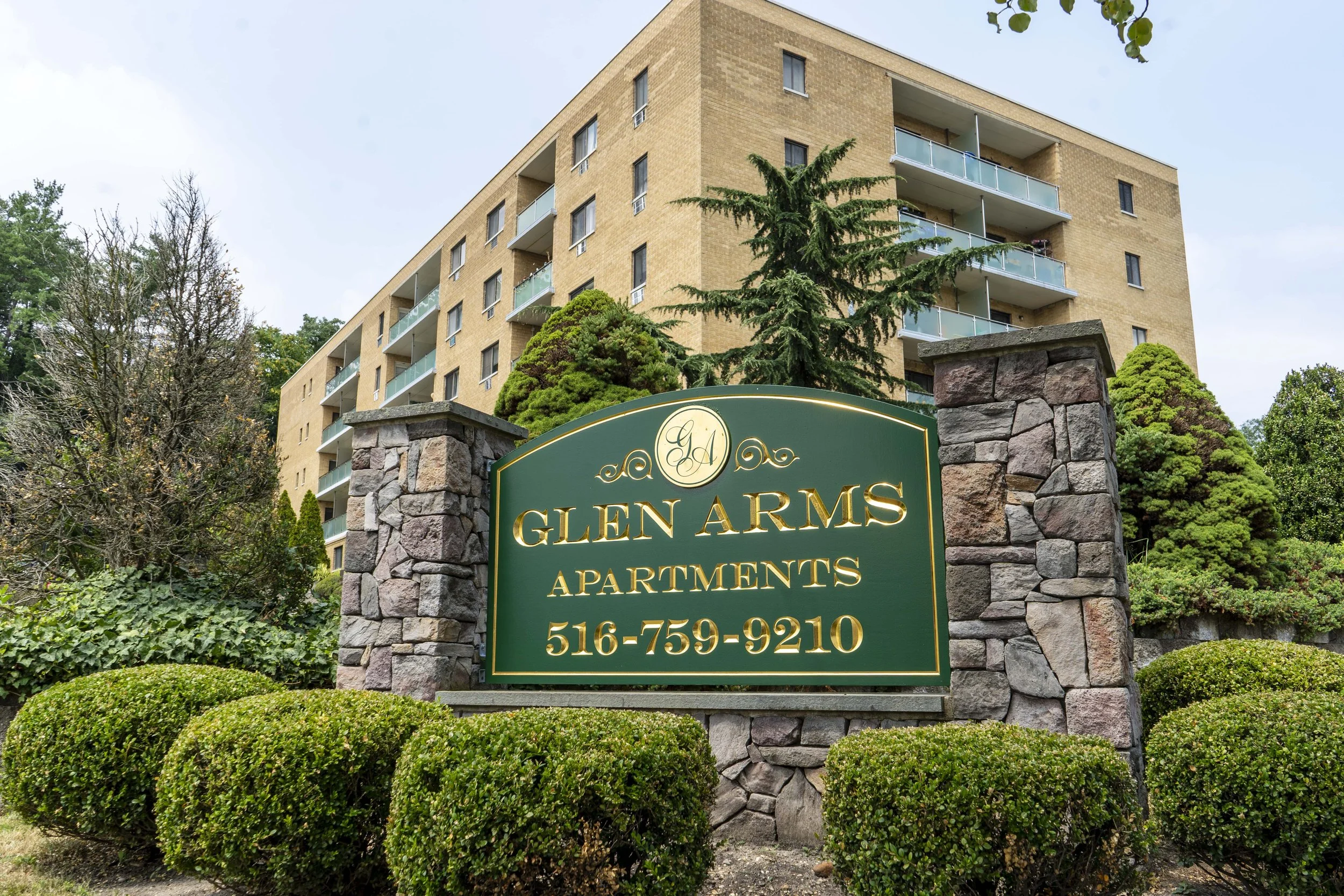 Glen Arms Apartments sign with contact number, a multi-story apartment building, trees, and bushes in the background.