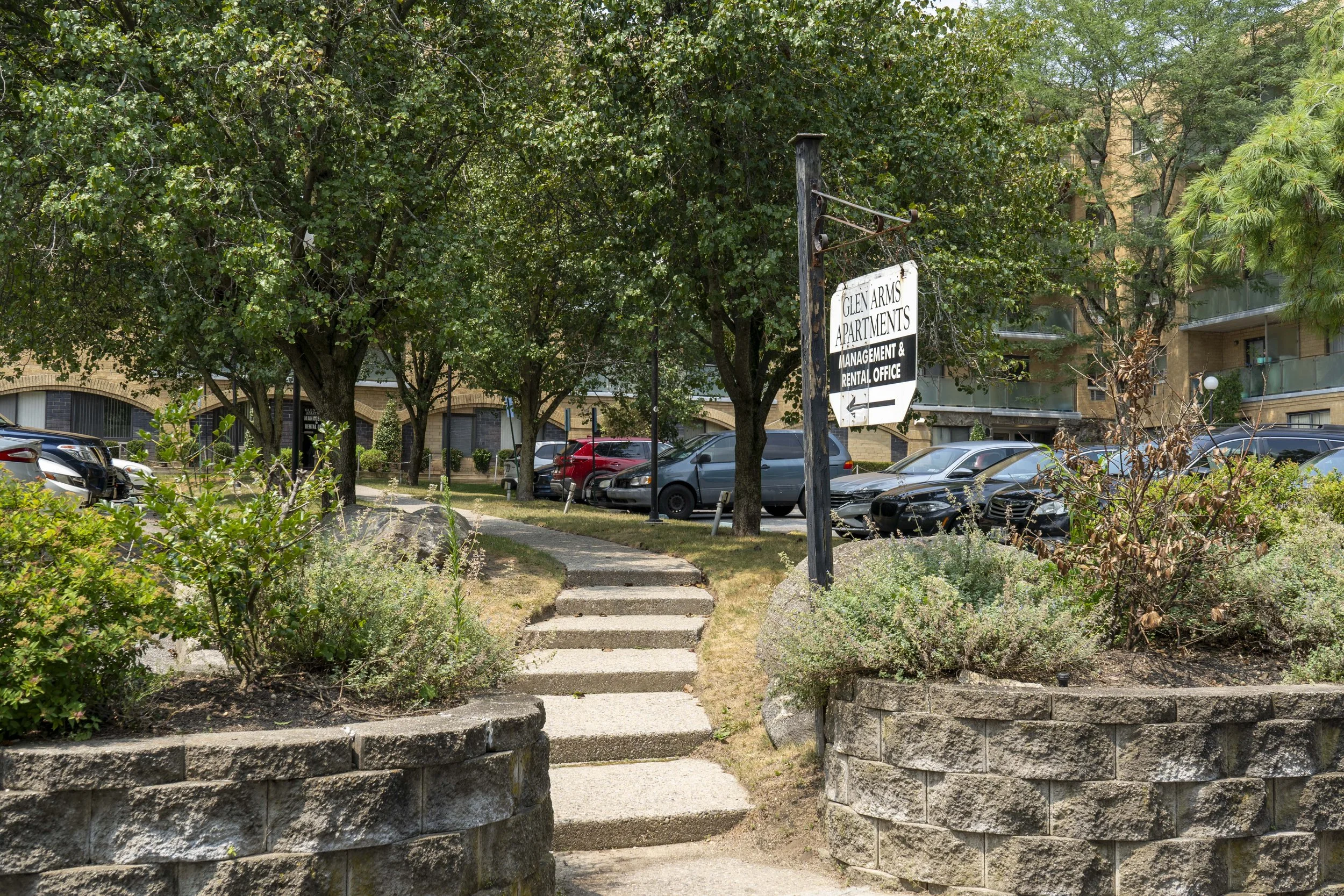 Sidewalk leading into a parking lot with trees and bushes, residential buildings in the background, and a sign indicating Glen Arms Apartments management and rental office.