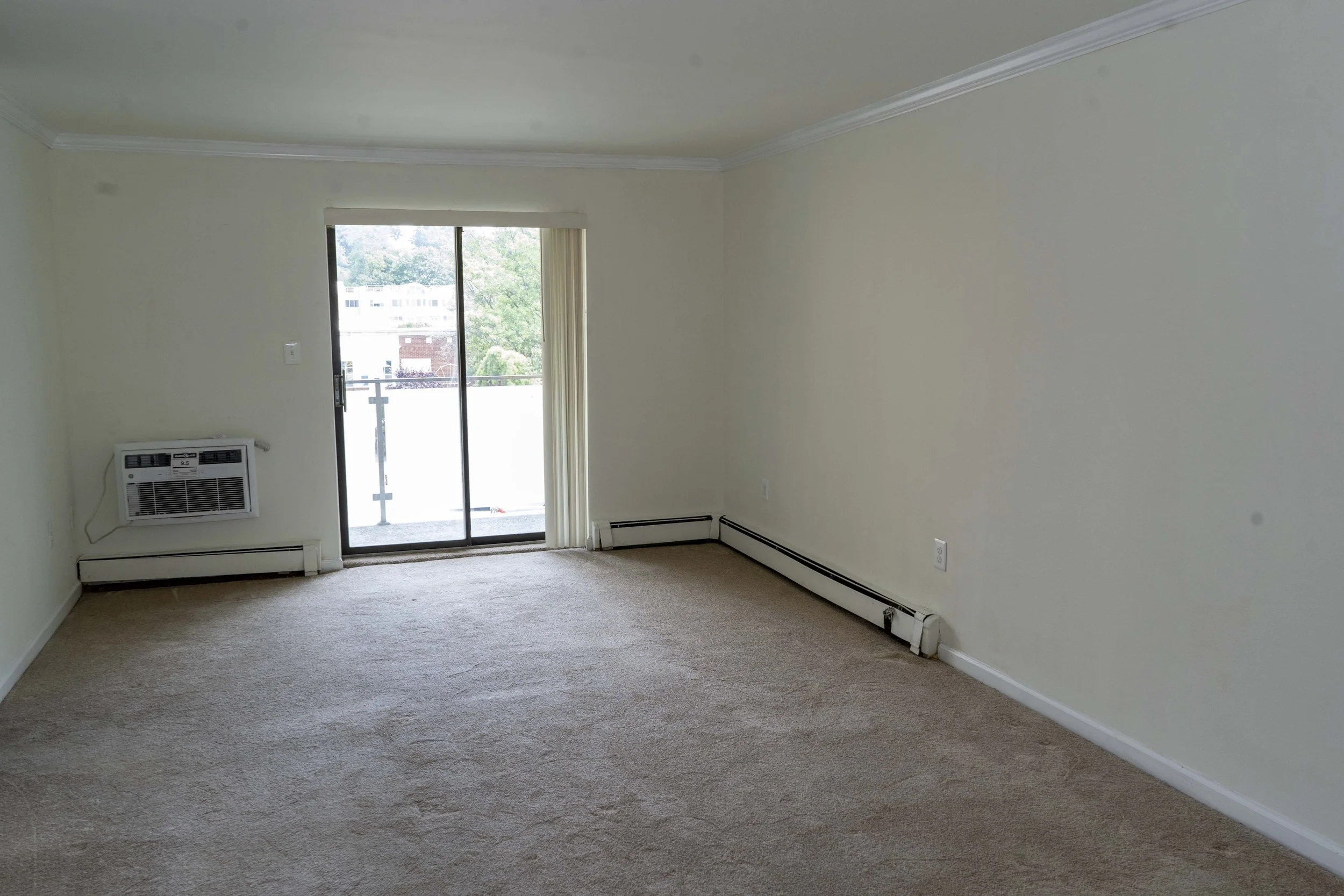 Empty living room with beige carpet, white walls, sliding glass door leading to a balcony, window air conditioner, and baseboard heaters.