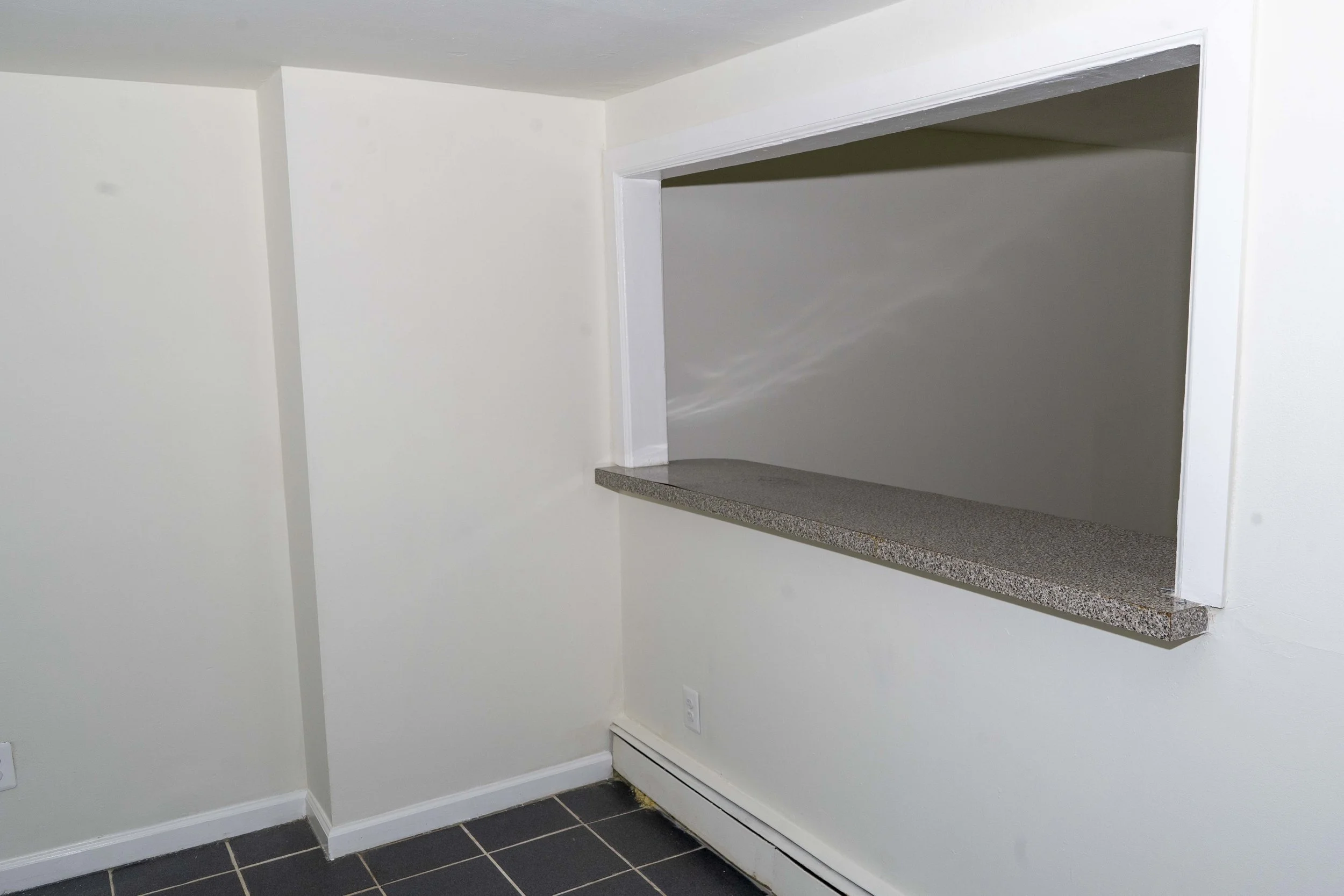 Empty wall with a square opening and a small ledge, likely a counter or shelf, in a room with black tiled flooring and white walls.