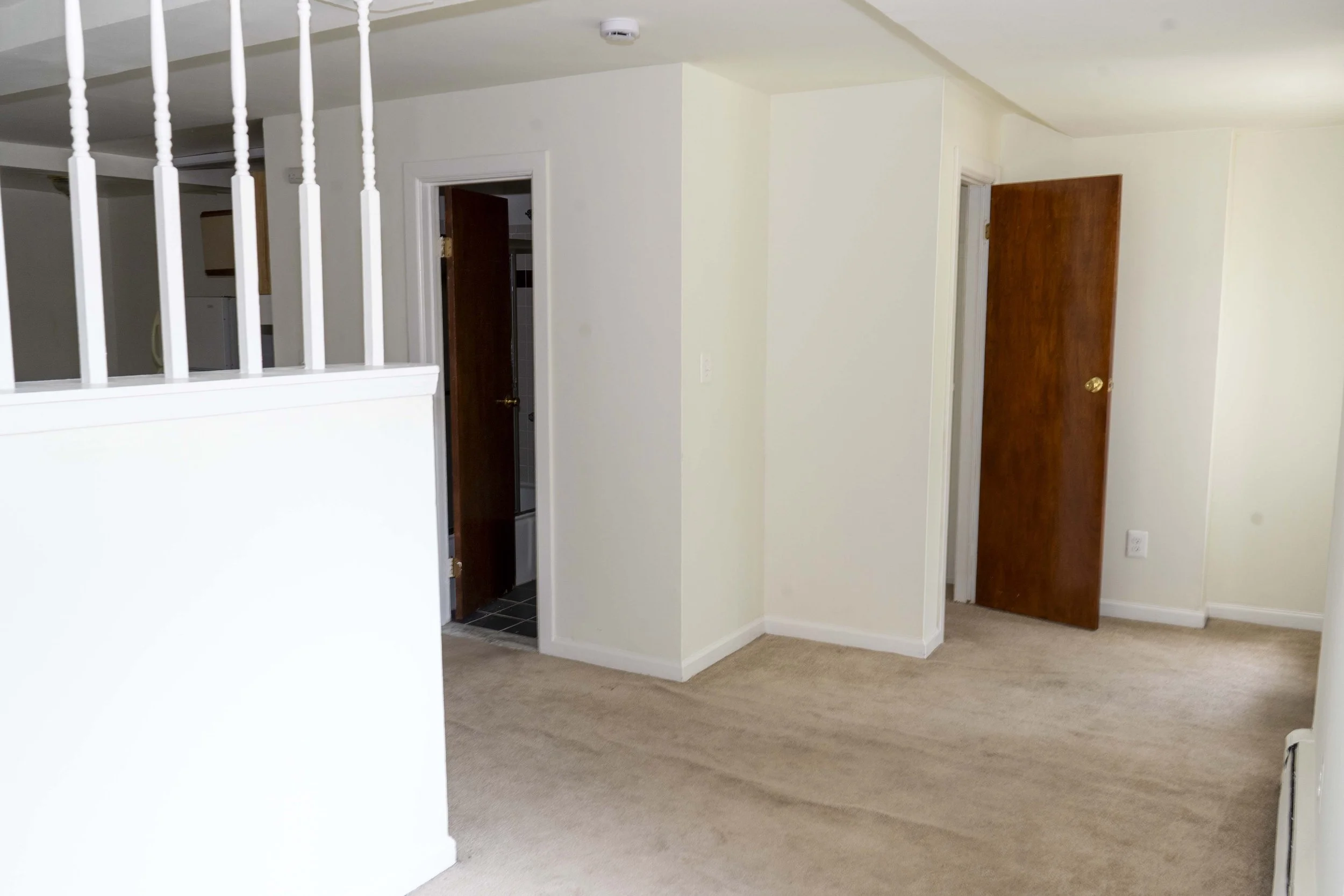 Empty room with beige carpet, white walls, and two closed wooden doors, one leading to a bathroom, with a partial view of a kitchen area to the left.