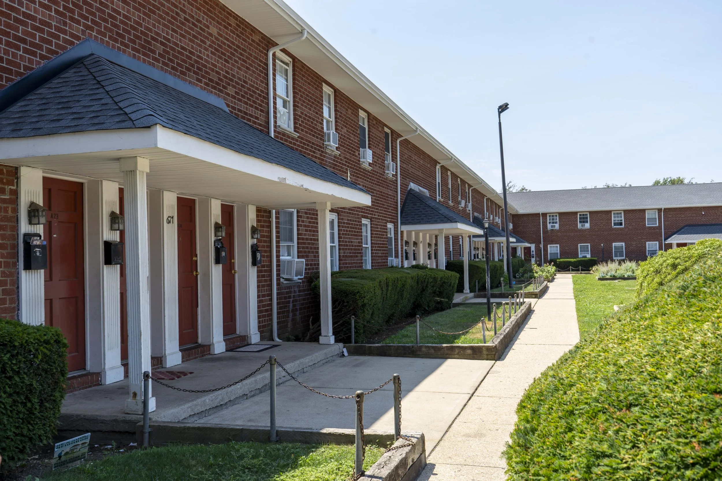 Row of brick apartment buildings with red doors, black mailboxes, small porches, and landscaped surroundings under a clear blue sky.