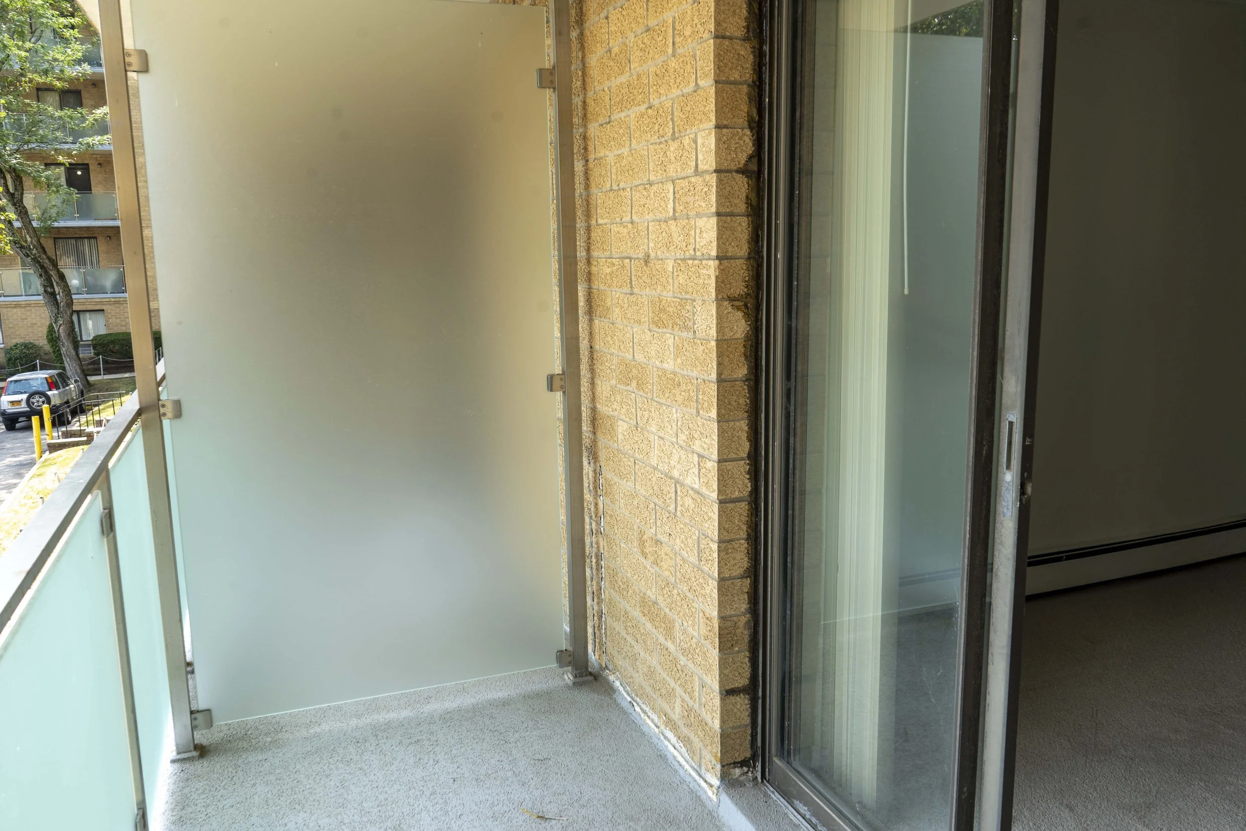Balcony with frosted glass privacy screen, metal railings, and a sliding glass door leading to an interior room, with a brick exterior wall.