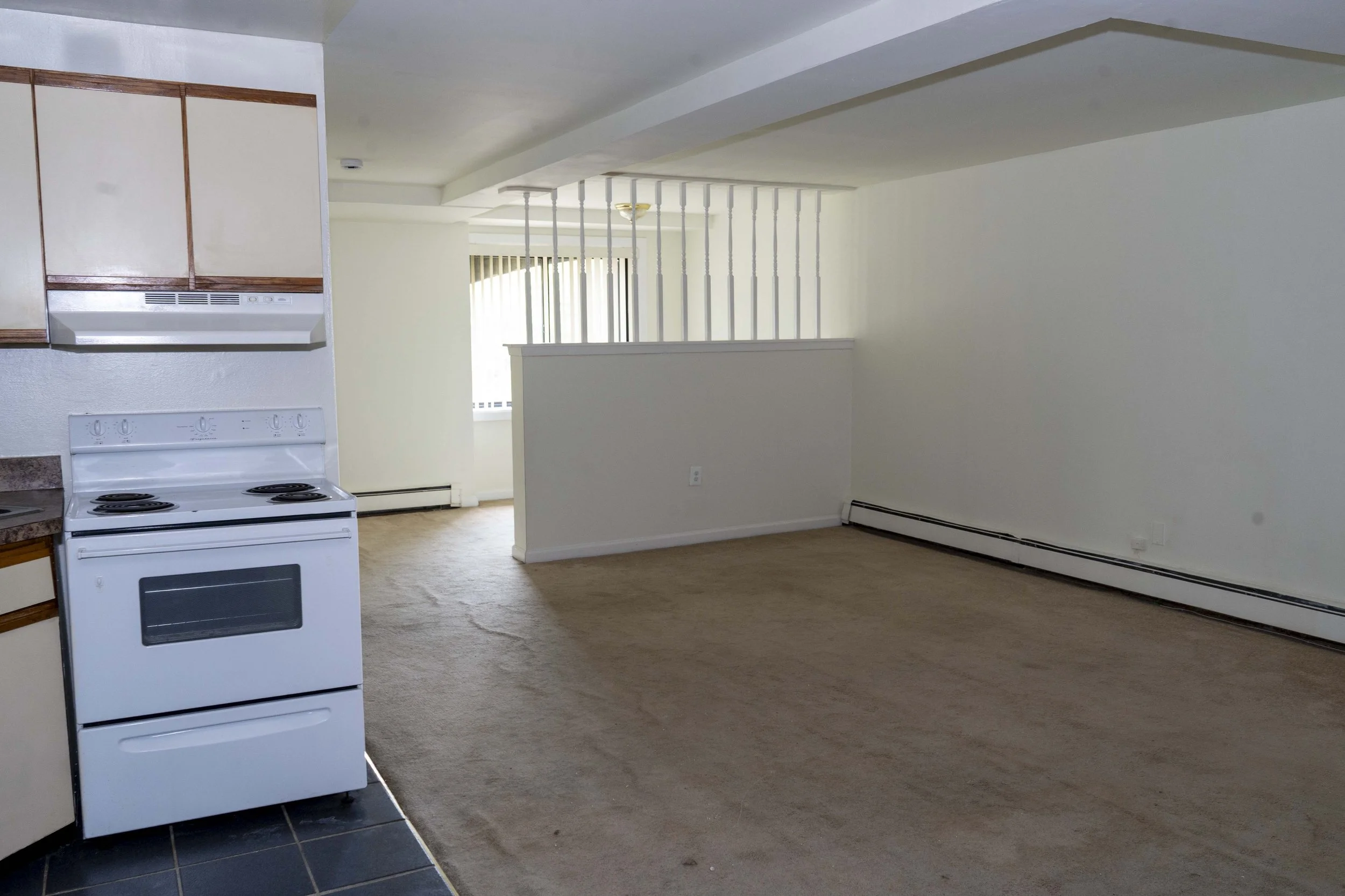 Empty living room and kitchen with white walls, beige carpet, and white stove, with a partial view of kitchen cabinets.