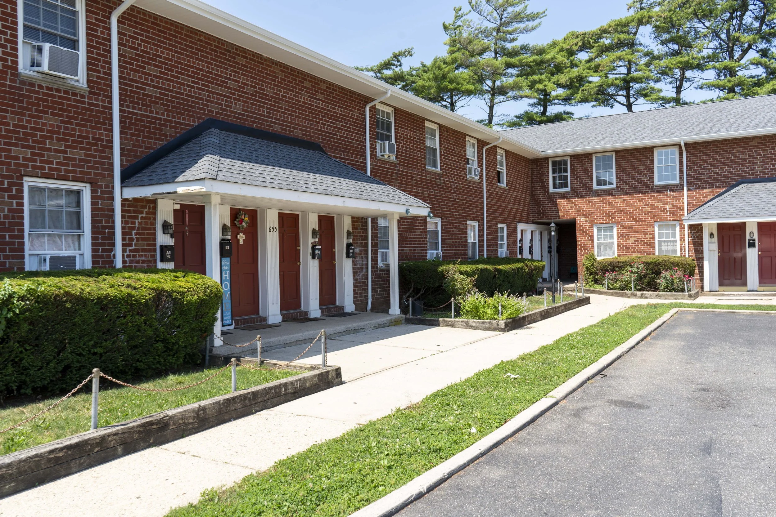 Exterior view of a red brick apartment building with multiple doors, small front gardens, and parked parking lot, under clear sky with pine trees in the background.