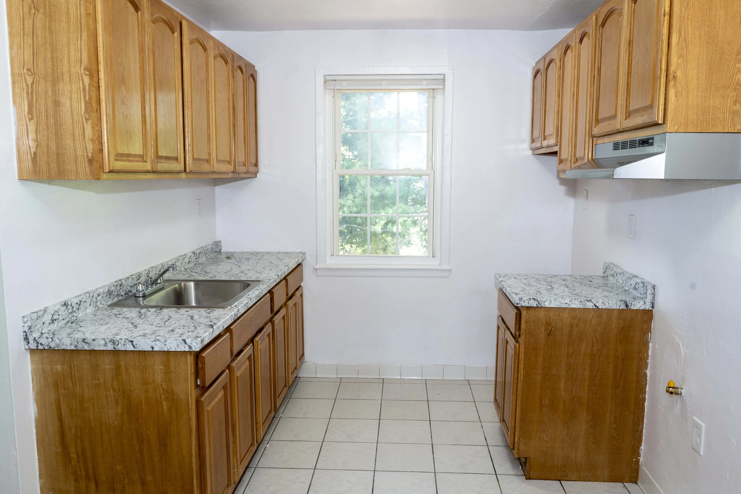A small kitchen with wooden cabinets, a granite countertop, a stainless steel sink, and a window showing trees outside.