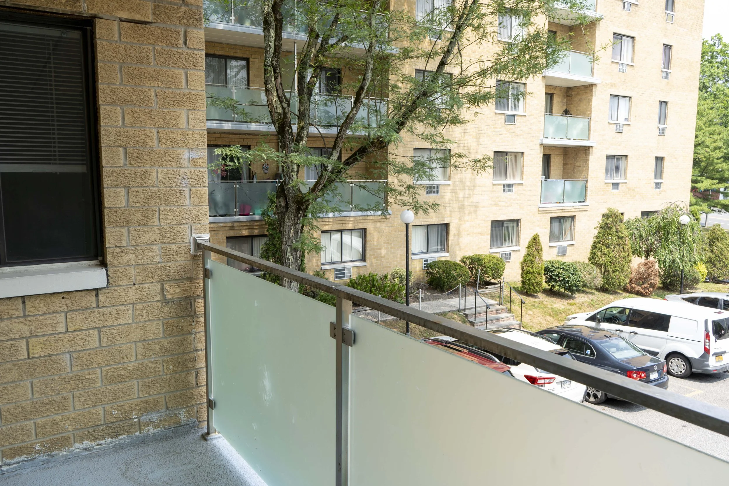 View from a balcony showing parked cars, a tree with green foliage, a landscaped area with shrubs, and a multi-story apartment building with brick walls and glass balconies.