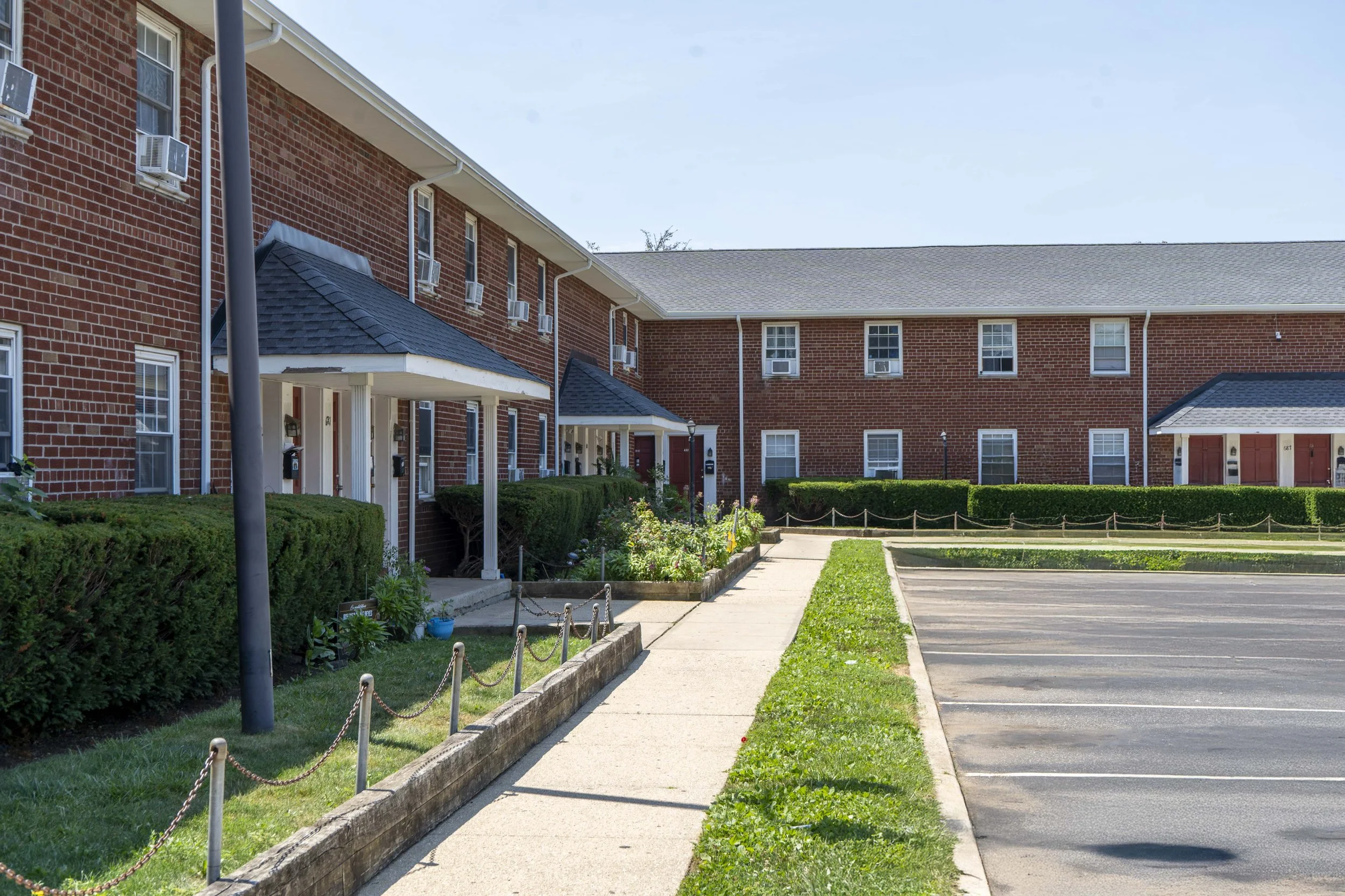 Exterior view of a red brick apartment complex with white porches, small shrubs, and an empty parking lot.