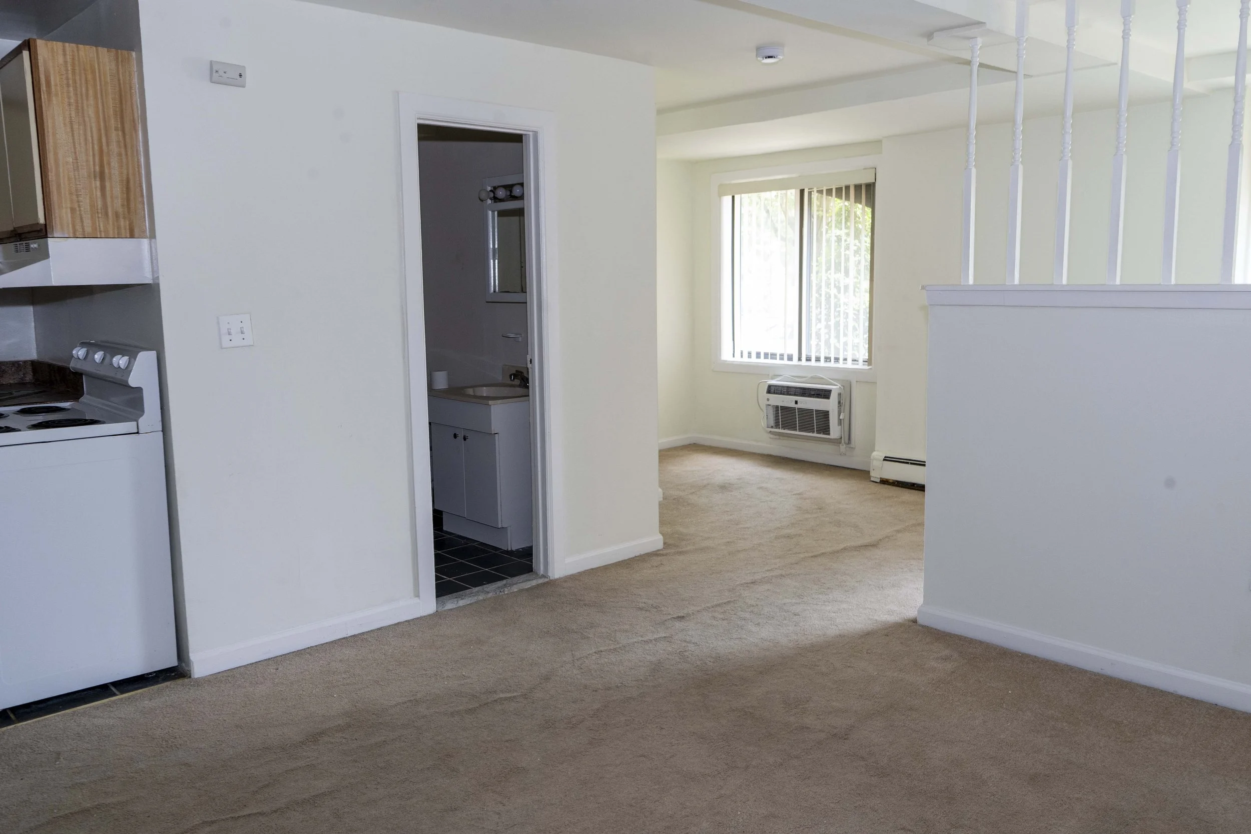 Empty living room and kitchen area with beige carpet, white walls, window air conditioning unit, and a bathroom entrance visible through an open door.