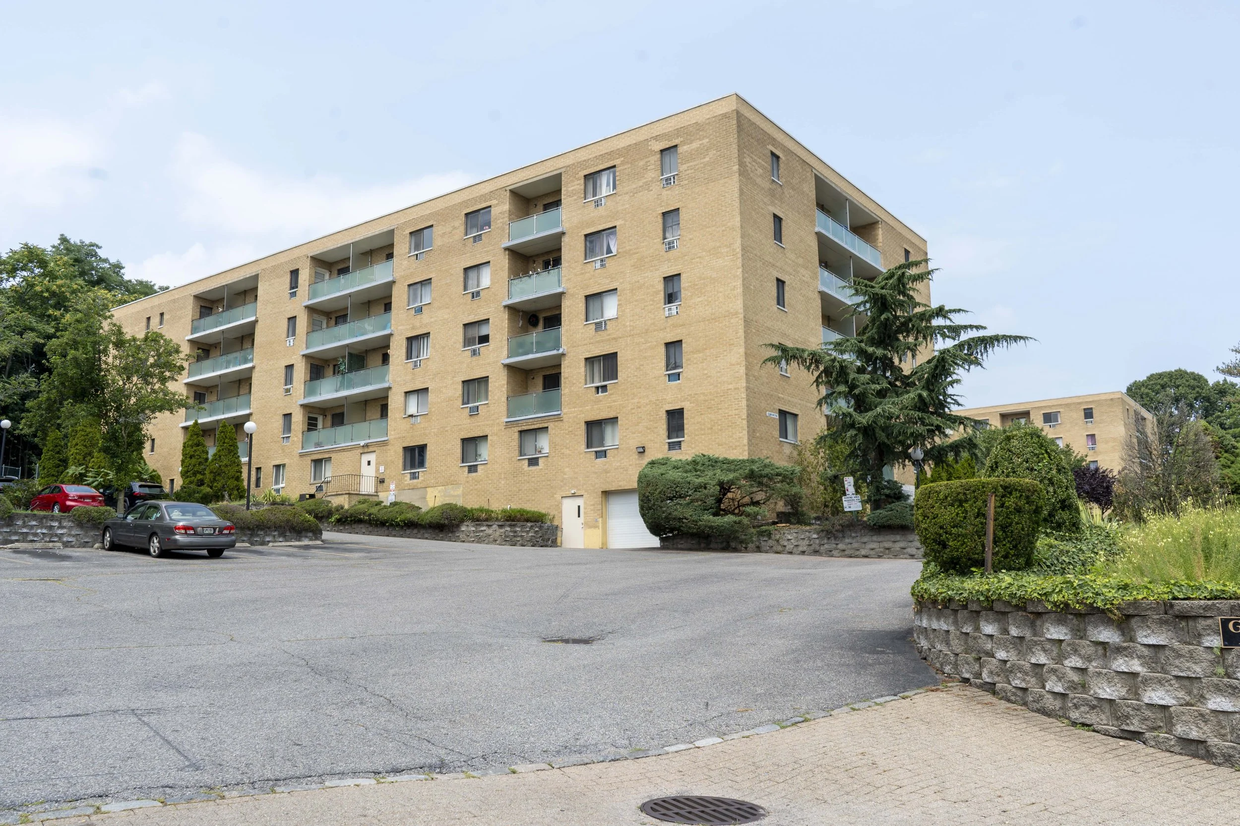 A multi-story residential apartment building with balconies and a parking lot in front. The building is made of tan bricks, surrounded by trees and greenery.