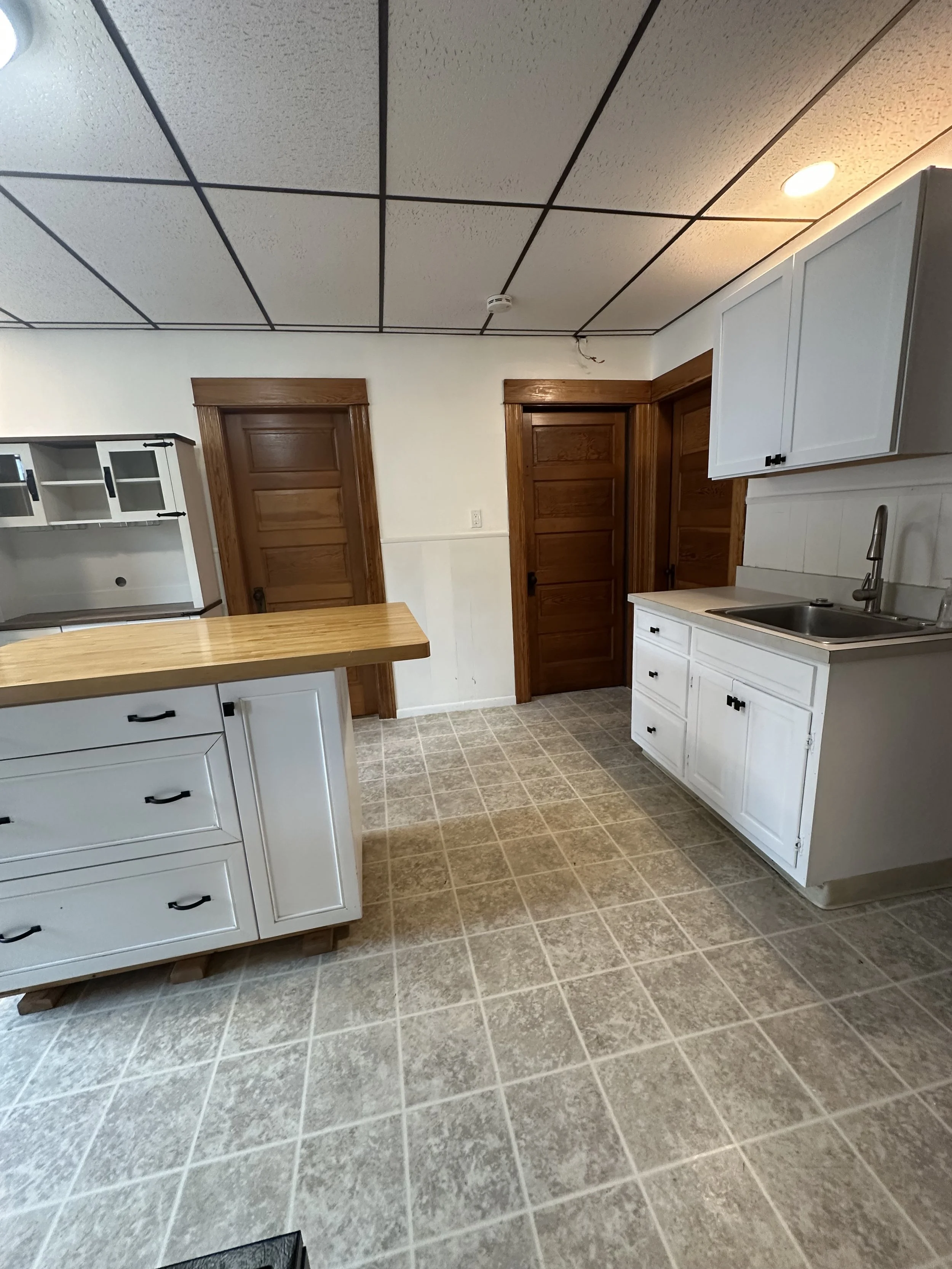 Empty kitchen with white cabinets, a wooden countertop island, two wooden doors, and tiled flooring.