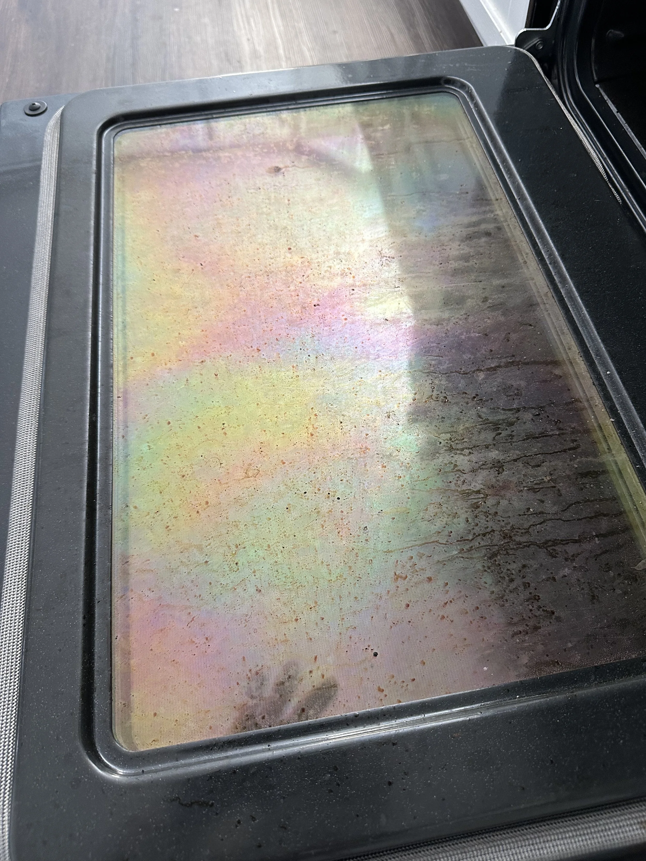 The glass on a stove or oven shows a greasy and stained surface with rainbow-colored marks.