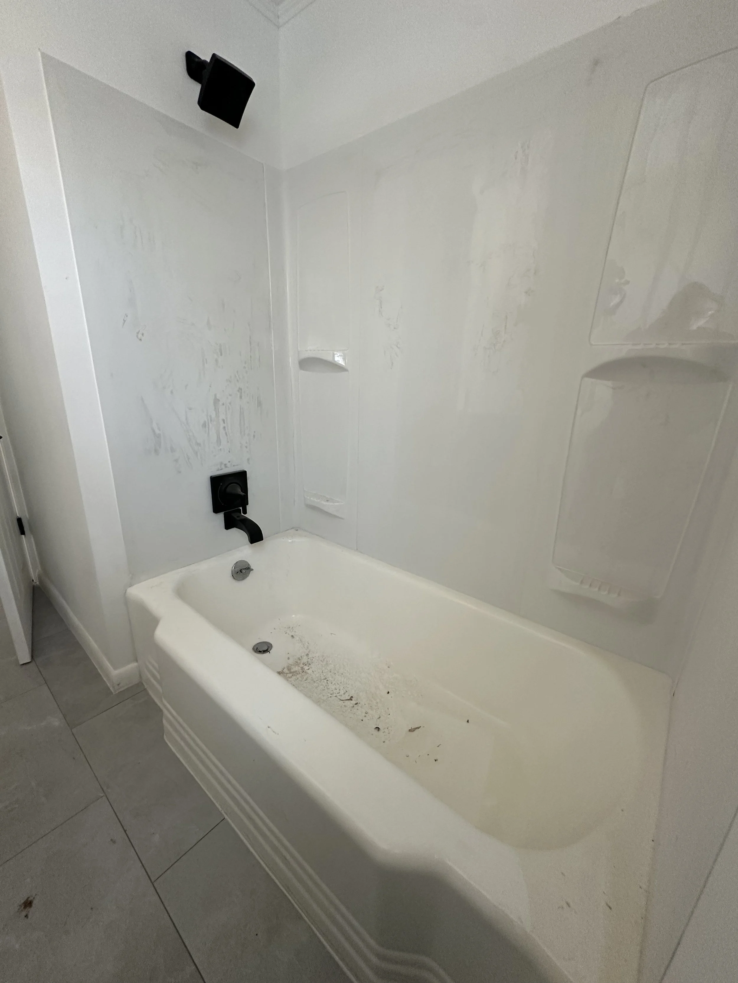 Cluttered bathtub with dirt, debris, and grime, with black faucet and showerhead, in a plain bathroom.