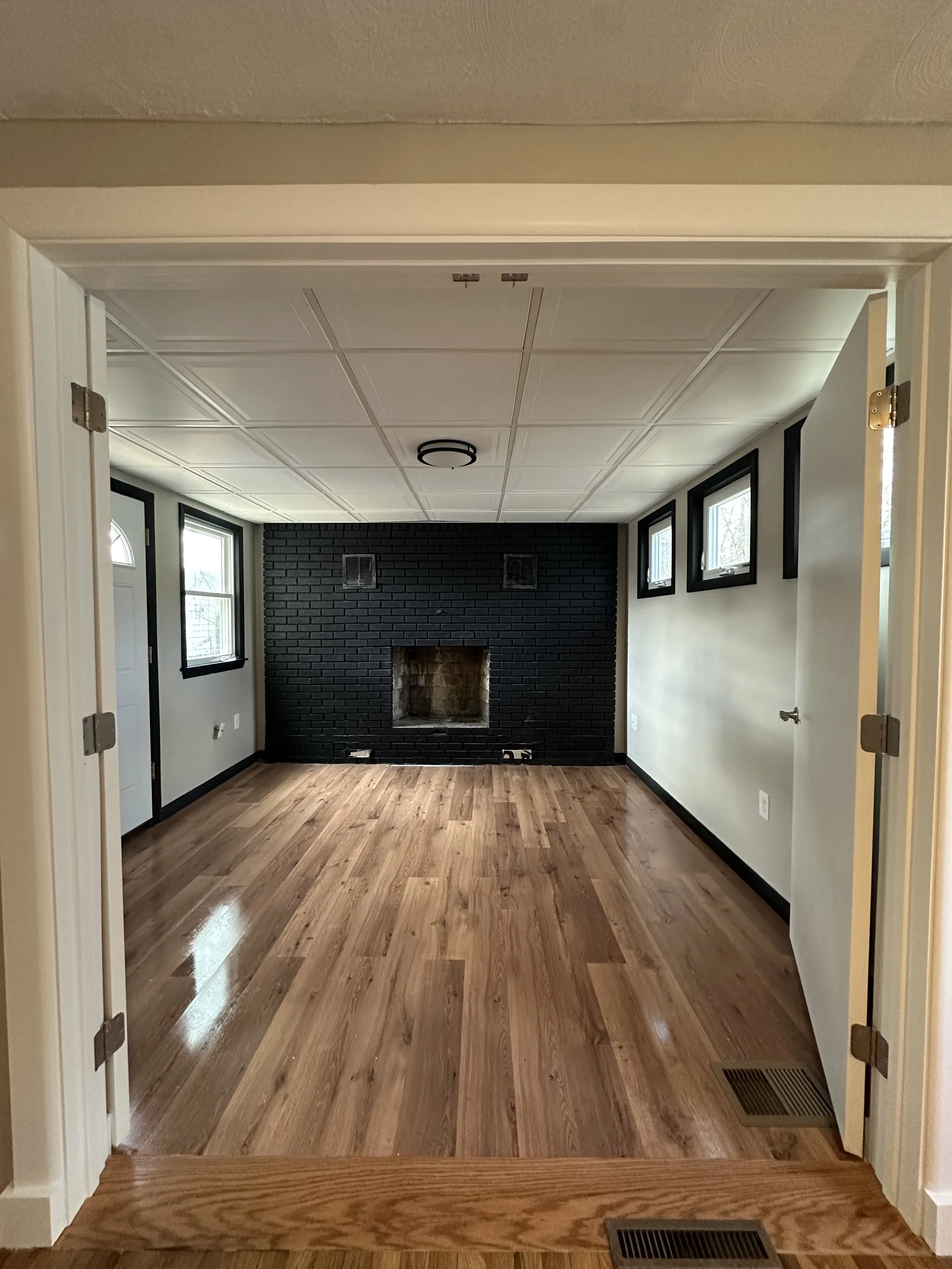 Empty living room with black brick fireplace, hardwood floors, and small rectangular windows on the right wall.