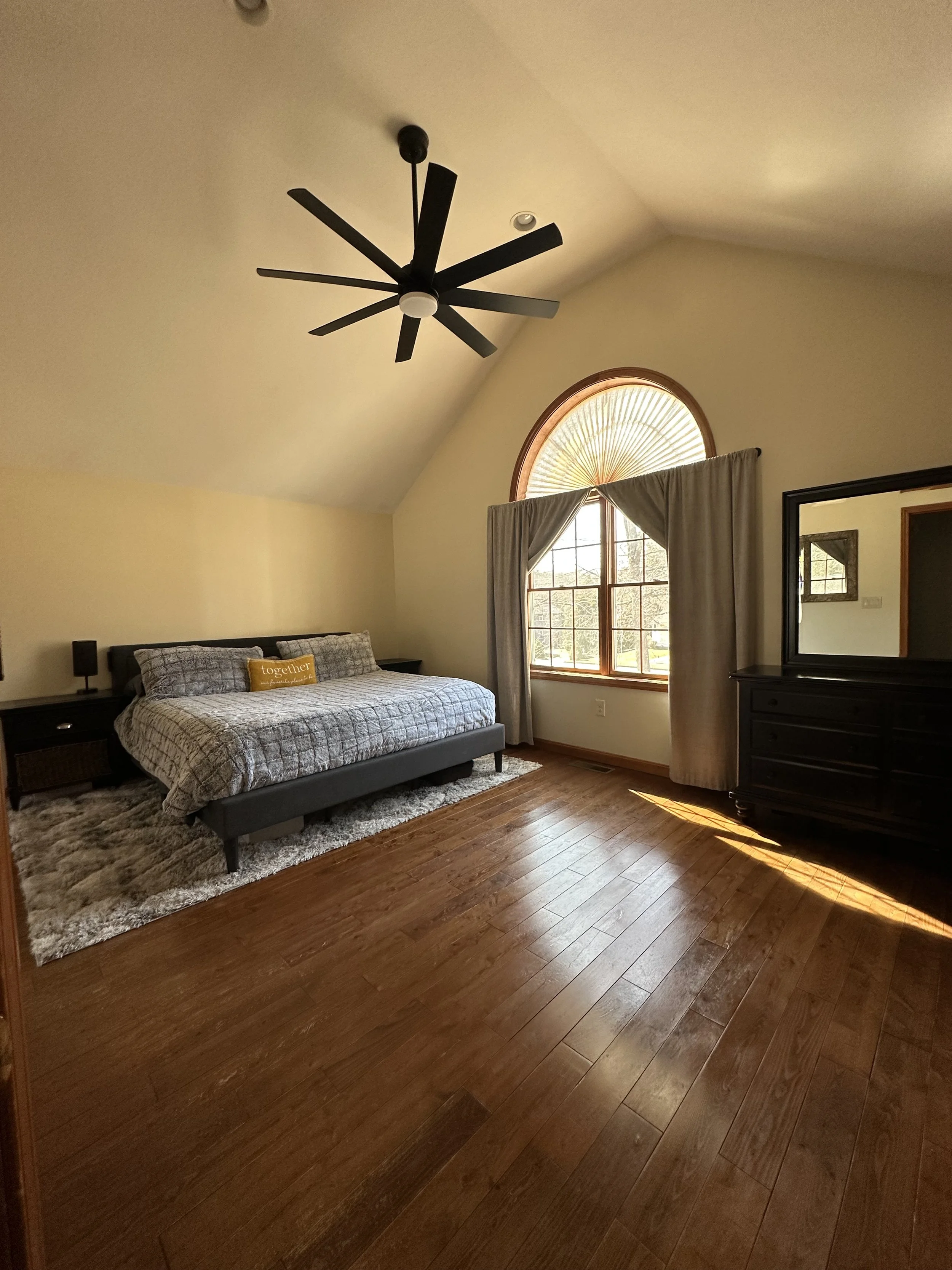 A spacious bedroom with hardwood floors, a large bed with gray bedding, a black dresser with a mirror, a large window with curtains, and a ceiling fan.