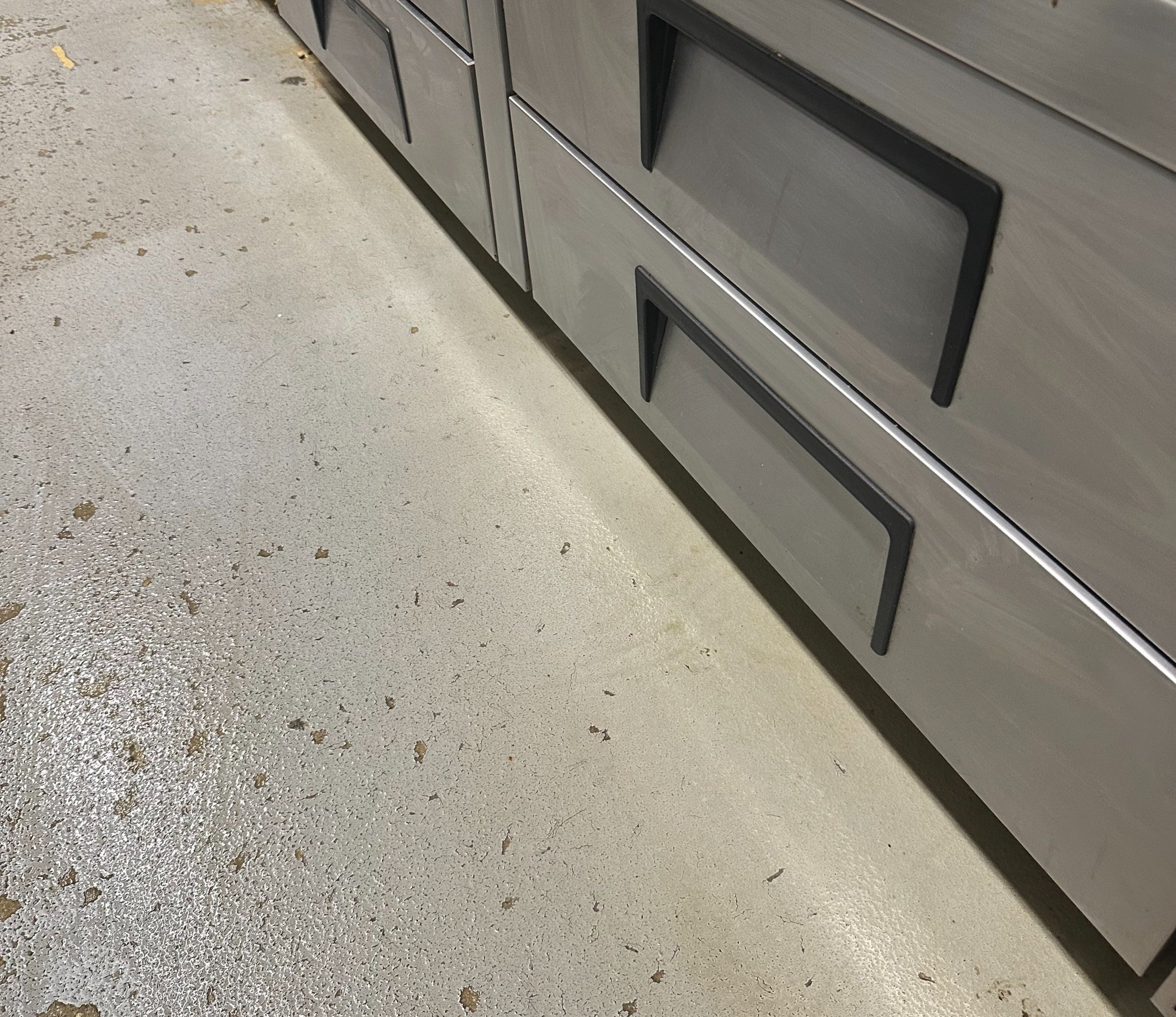 Close-up of stainless steel kitchen cabinets with black handles and a light-colored concrete floor.