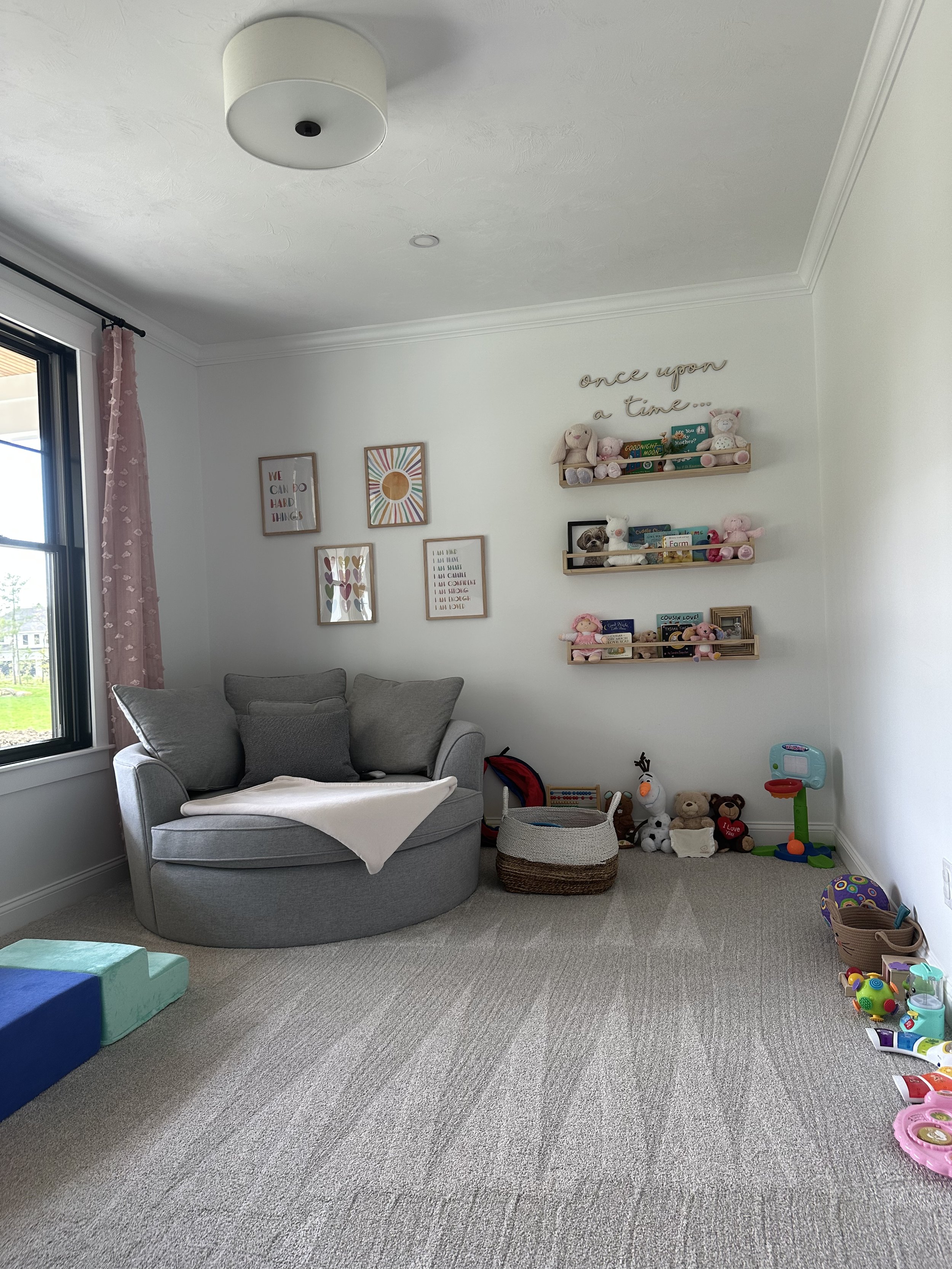 Living room with a gray sofa, wall art, and shelves filled with stuffed animals and toys, large window with pink curtains, toys on the carpeted floor.