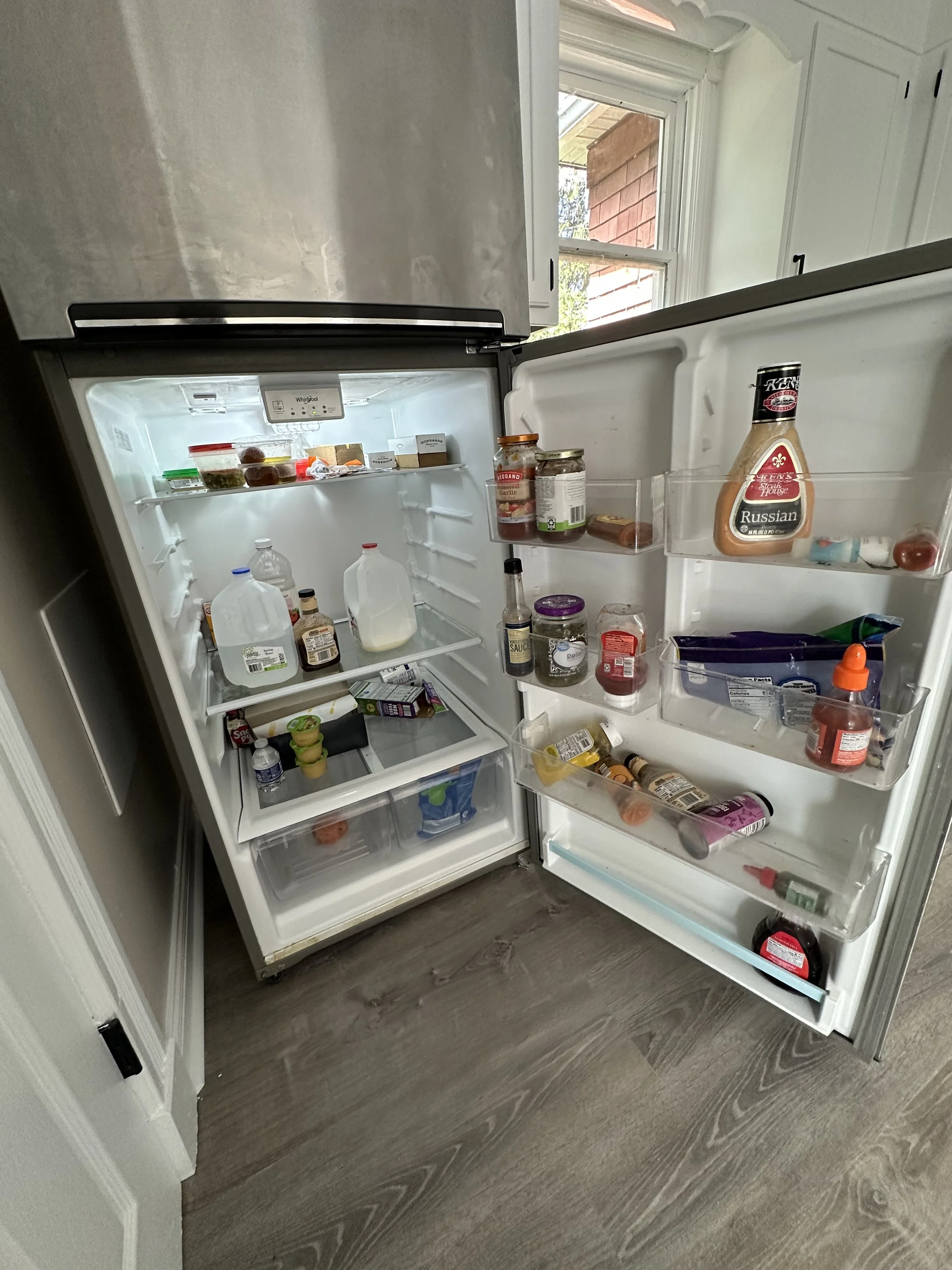 Open refrigerator door with various bottles, jars, and food containers inside, on a wood floor next to a window.
