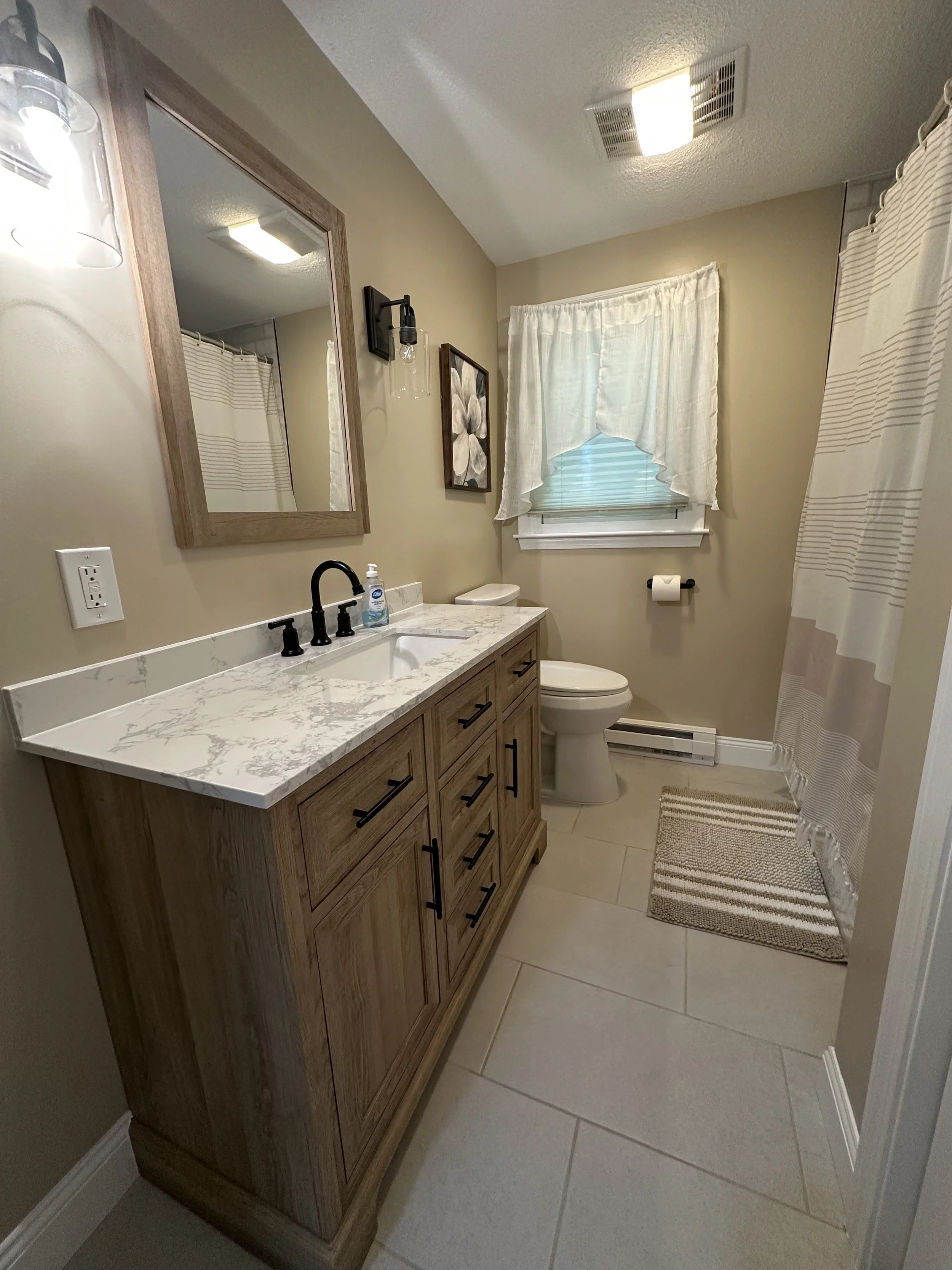 A small bathroom with a wooden vanity with a marble countertop, a white sink, and black fixtures. There is a mirror above the sink, a wall-mounted light fixture, a framed picture on the wall, a window with white curtains, a toilet, a toilet paper rol