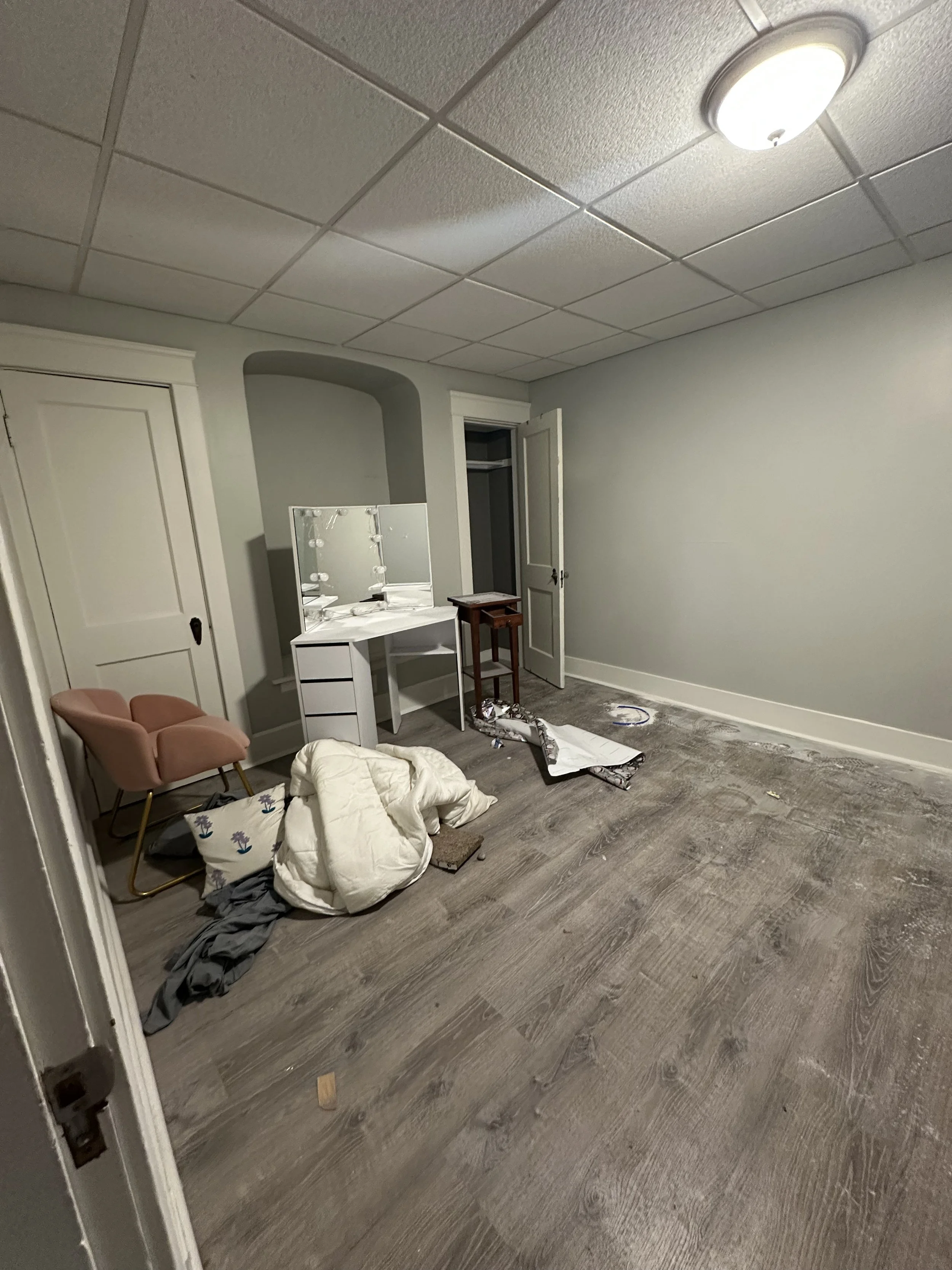 Empty room with light gray walls, a ceiling light, a pink armchair, a white vanity table with a mirror, a small wooden table, and scattered objects and debris on the wooden floor.