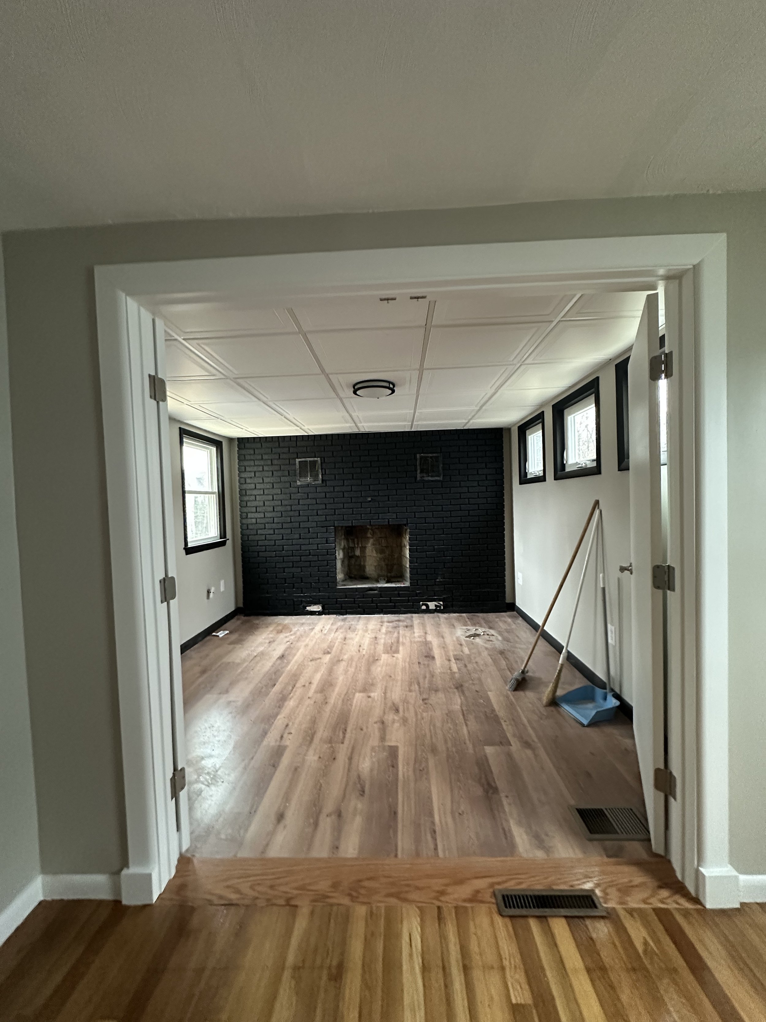 Living room with four windows, black brick fireplace, hardwood floor, and cleaning tools leaning against the wall.