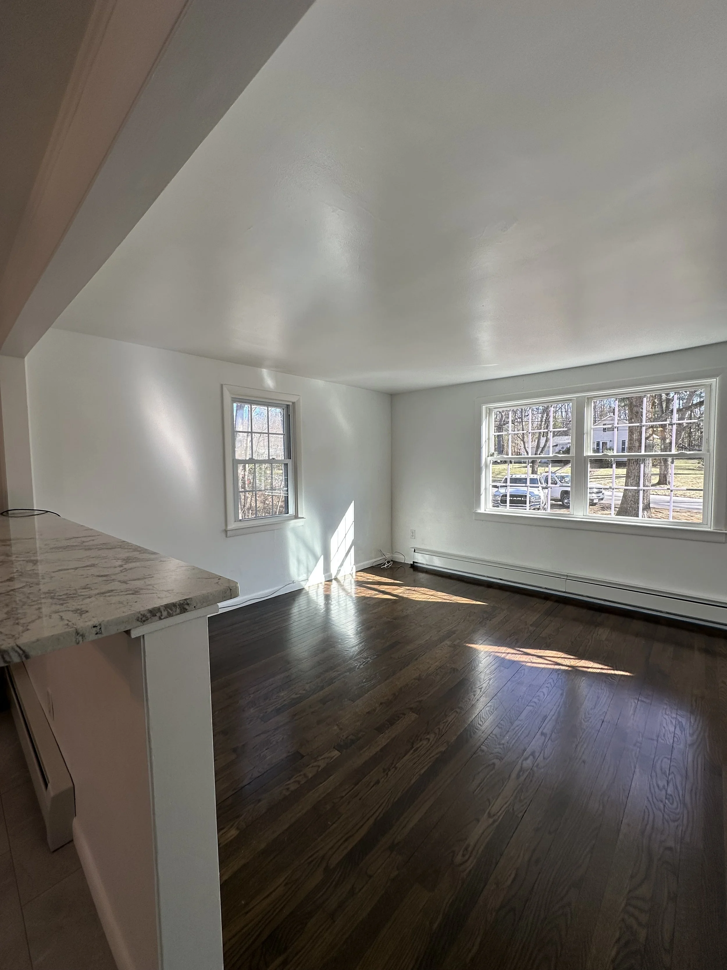 Empty living room with large windows, hardwood floors, white walls, and sunlight coming through the windows.