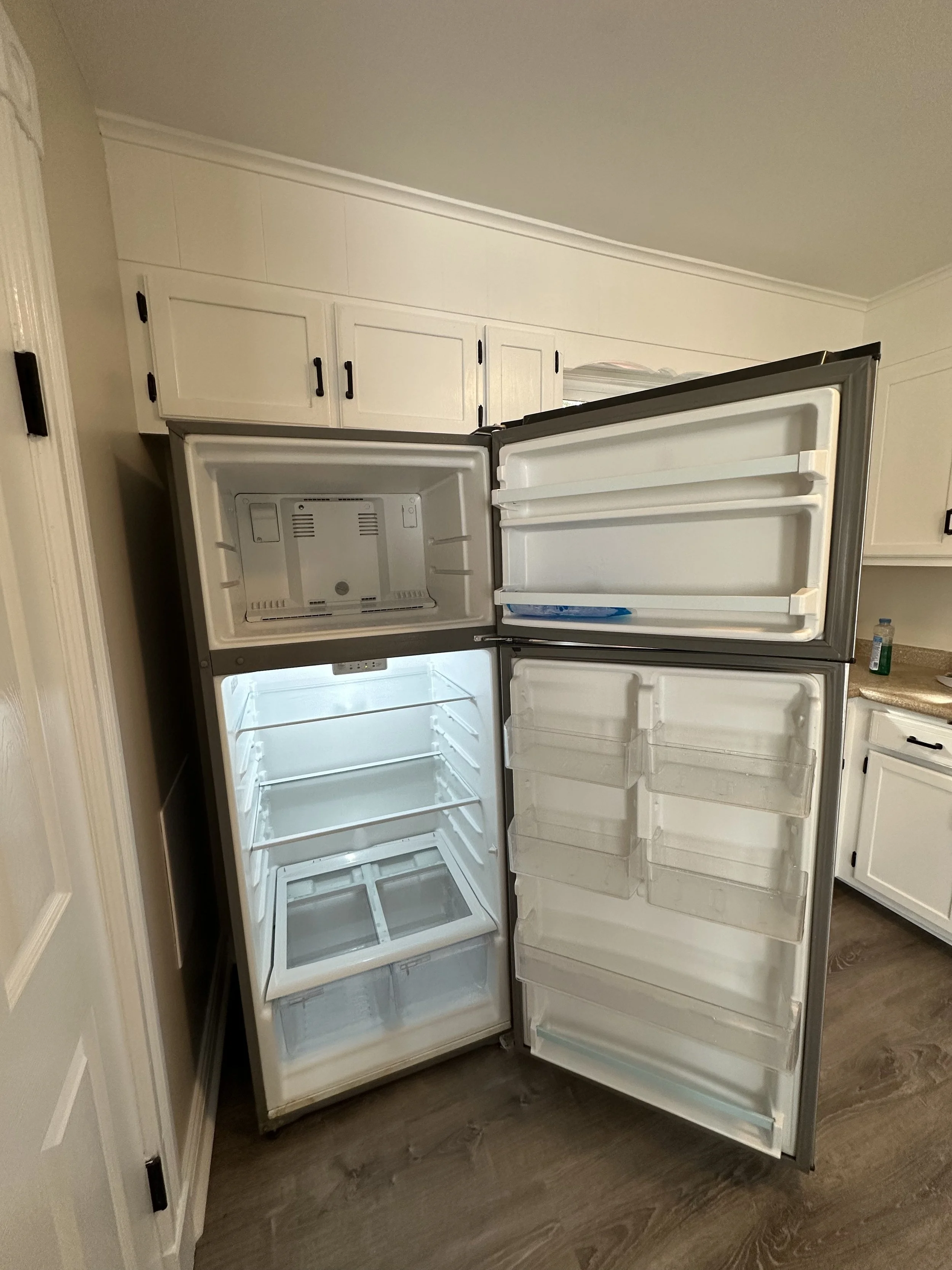 Open empty refrigerator with shelves inside, in a kitchen with white cabinets and wood flooring.
