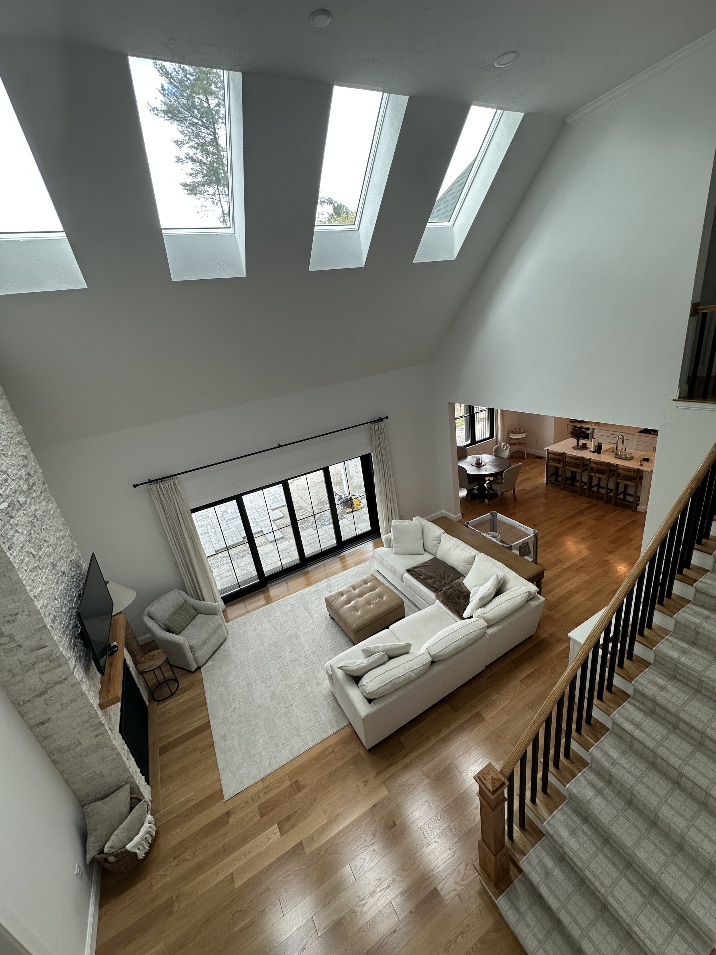 Living room with high ceiling, three skylights, a white sectional sofa, a beige tufted ottoman, gray armchair, large sliding glass doors with curtains, hardwood floors, and a staircase with a carpeted runner.