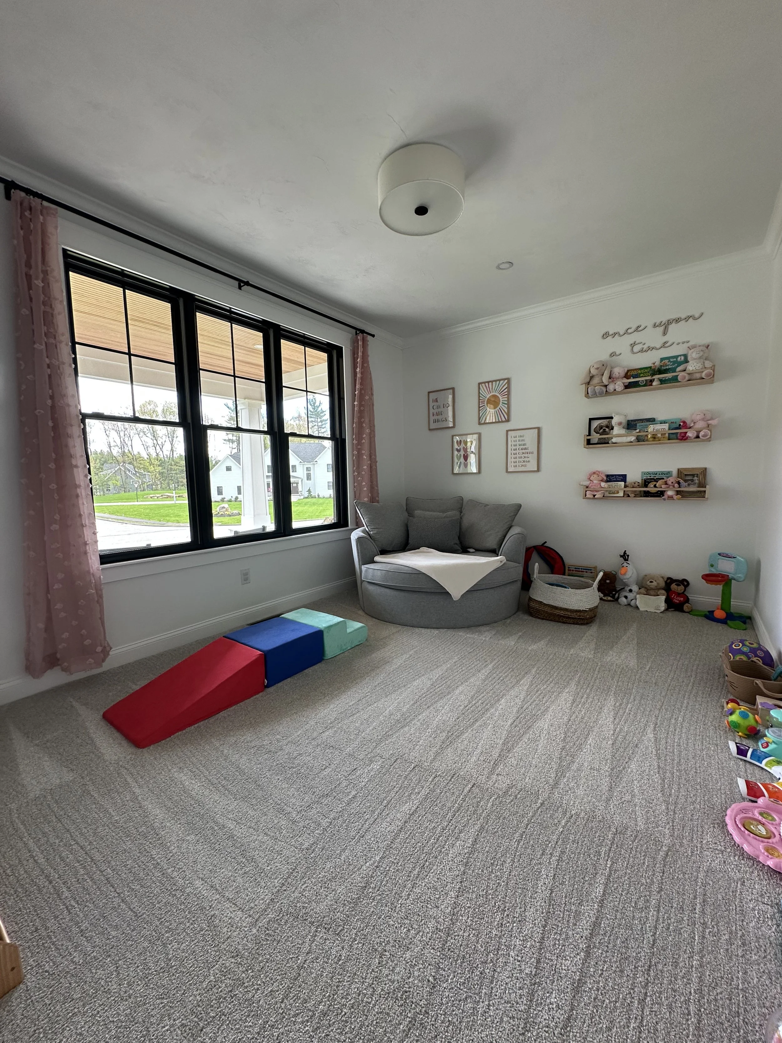A child's playroom with large windows, pink curtains, and stuffed animals on the wall shelves. There are colorful foam blocks, toys, and a small basketball hoop on the right side. The room has a gray round chair with pillows and framed wall art with 