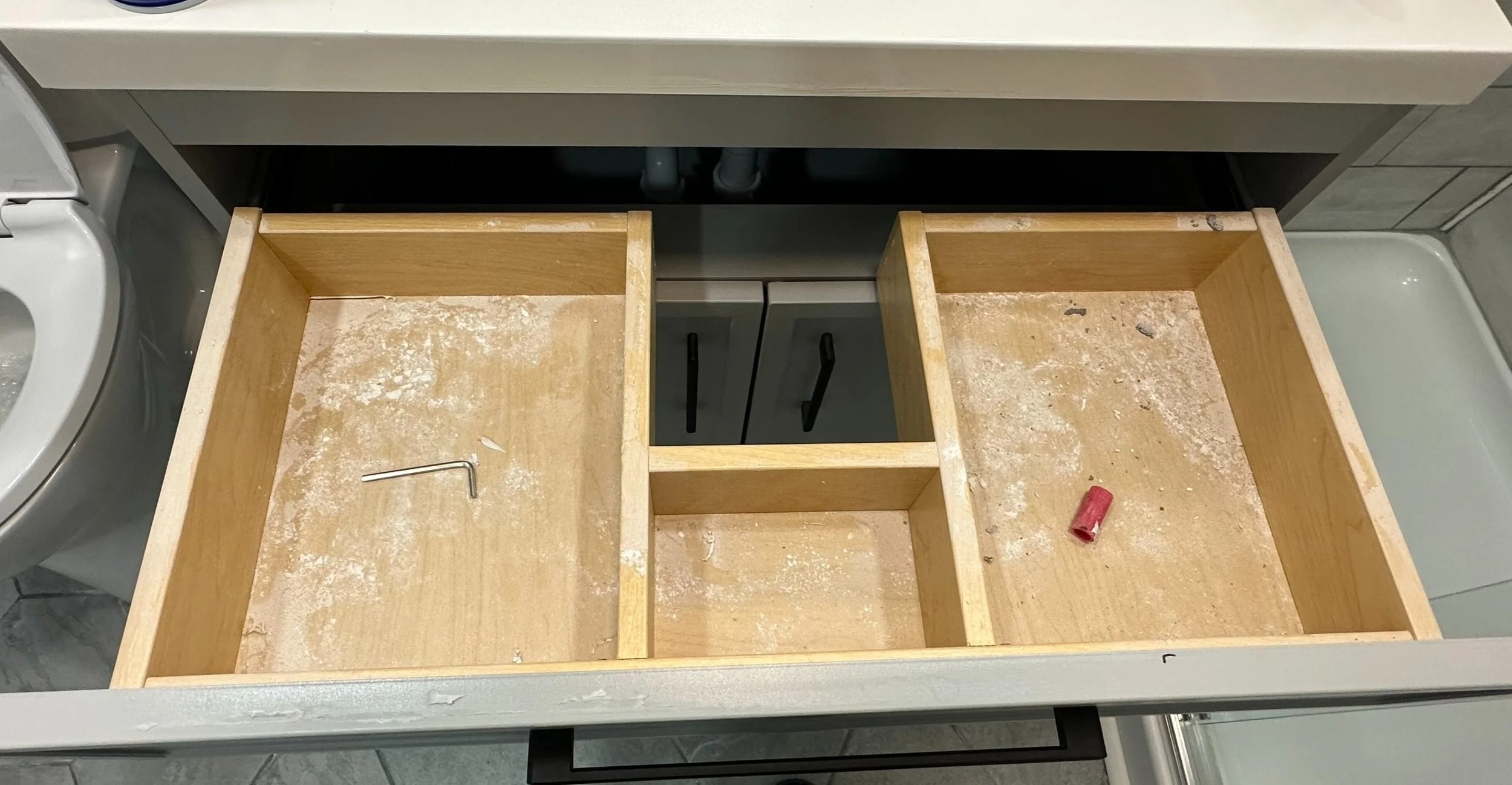 Open kitchen drawer being repaired, with dust and debris inside, a small Allen wrench and red socket in the drawer.
