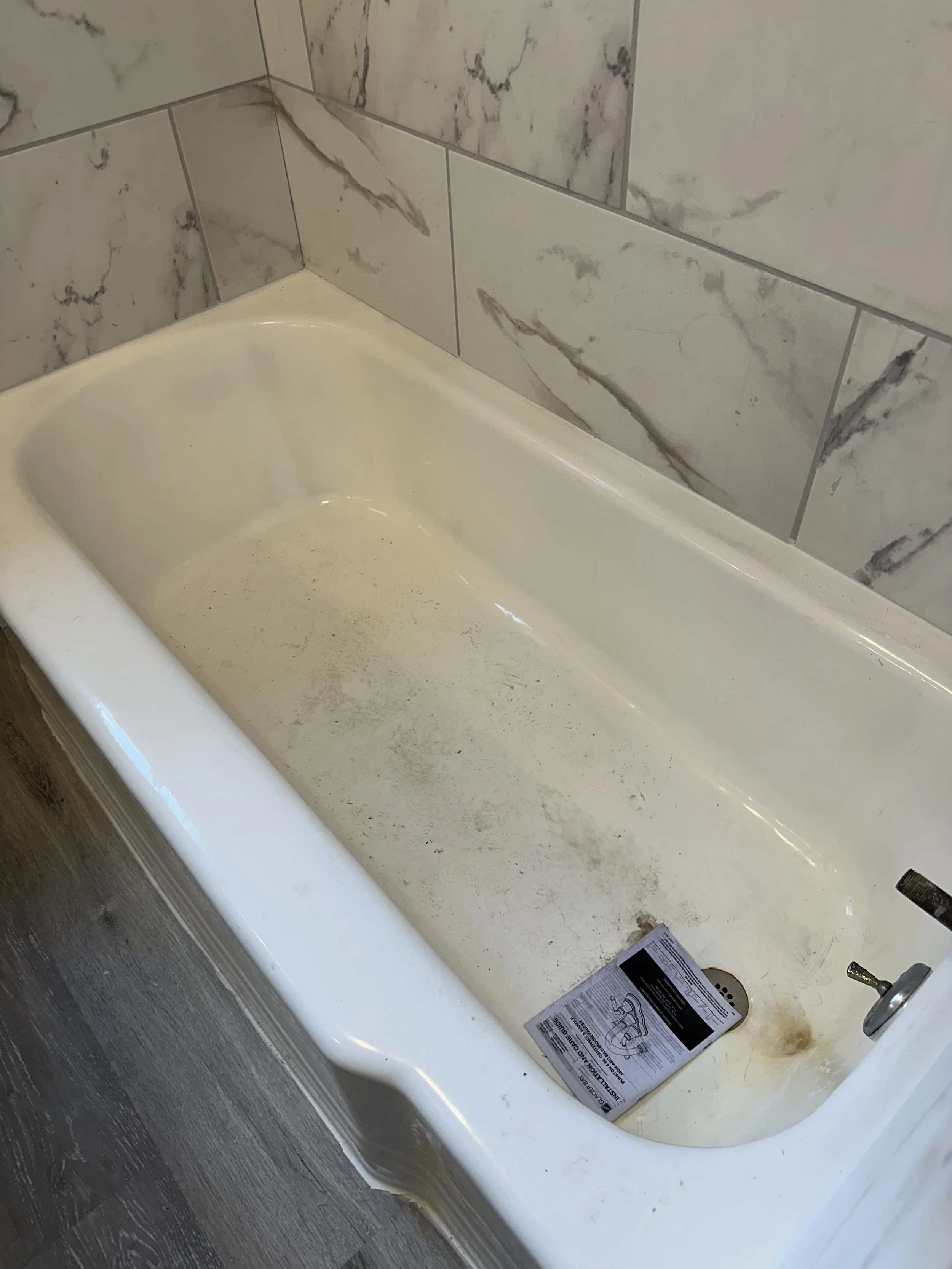 Dirty bathtub with soap scum and debris, located in a bathroom with marble tiles on the wall, and a newspaper or instruction sheet inside.