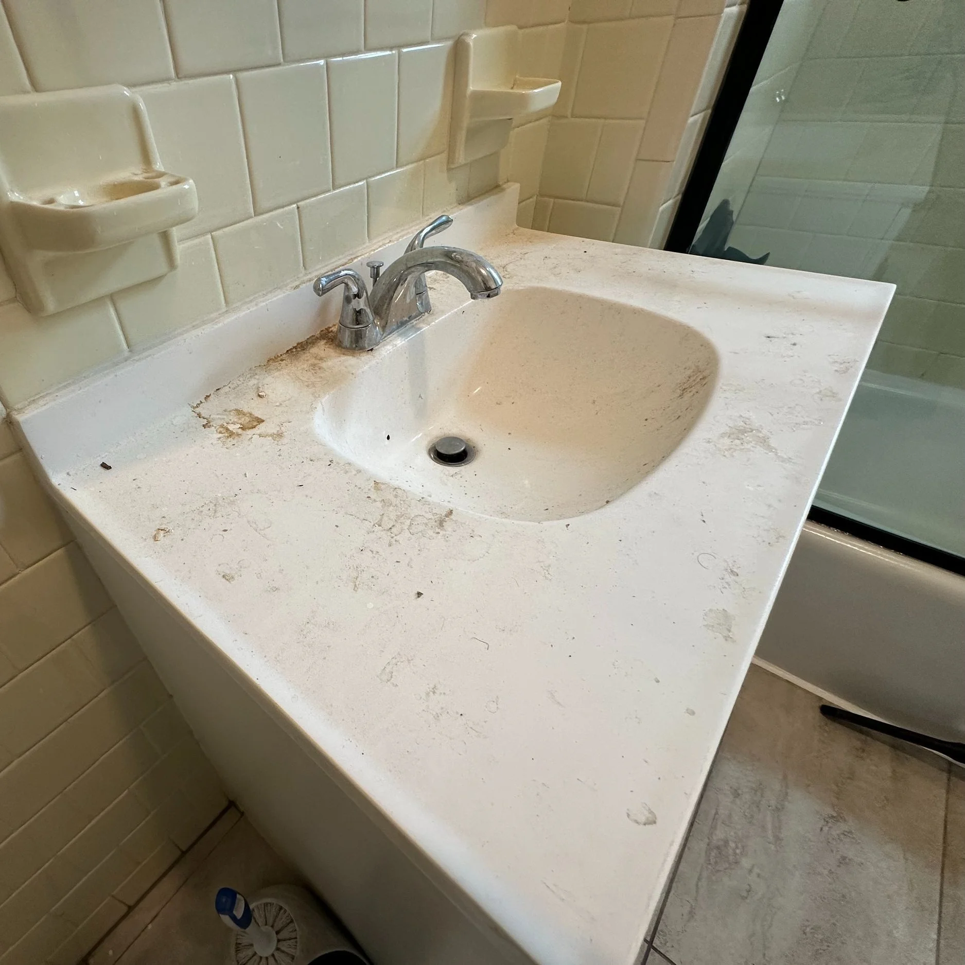 Dirty bathroom sink with stains, grime, and debris on a white countertop, with yellow tiled walls and a shower visible in the background.