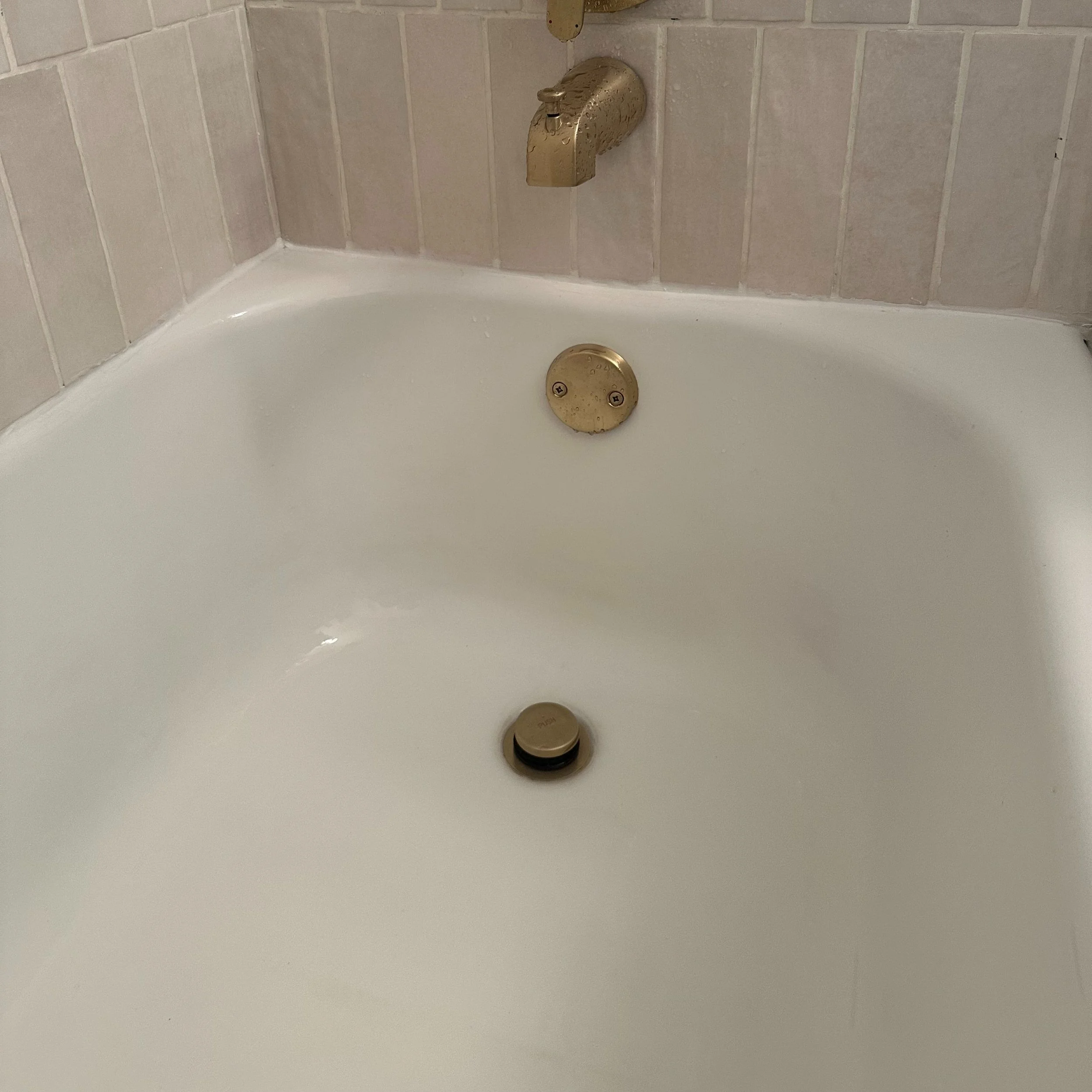 Empty bathtub with gold-colored drain and faucet in a bathroom with beige tiled walls.