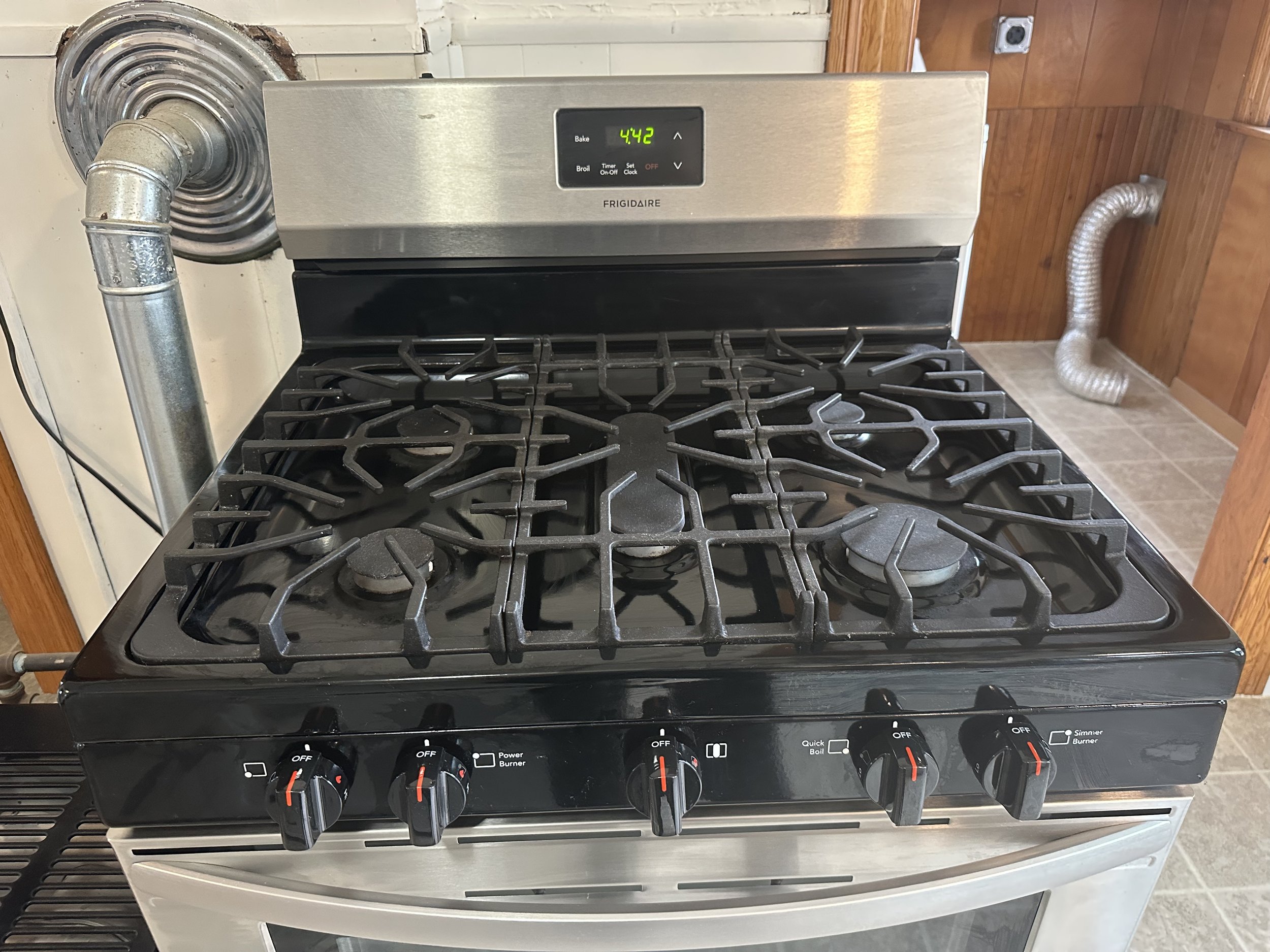 A stainless steel stove with four gas burners and black control knobs, with a digital clock displaying 4:42, situated in a kitchen with wood-paneled walls and tiled flooring.