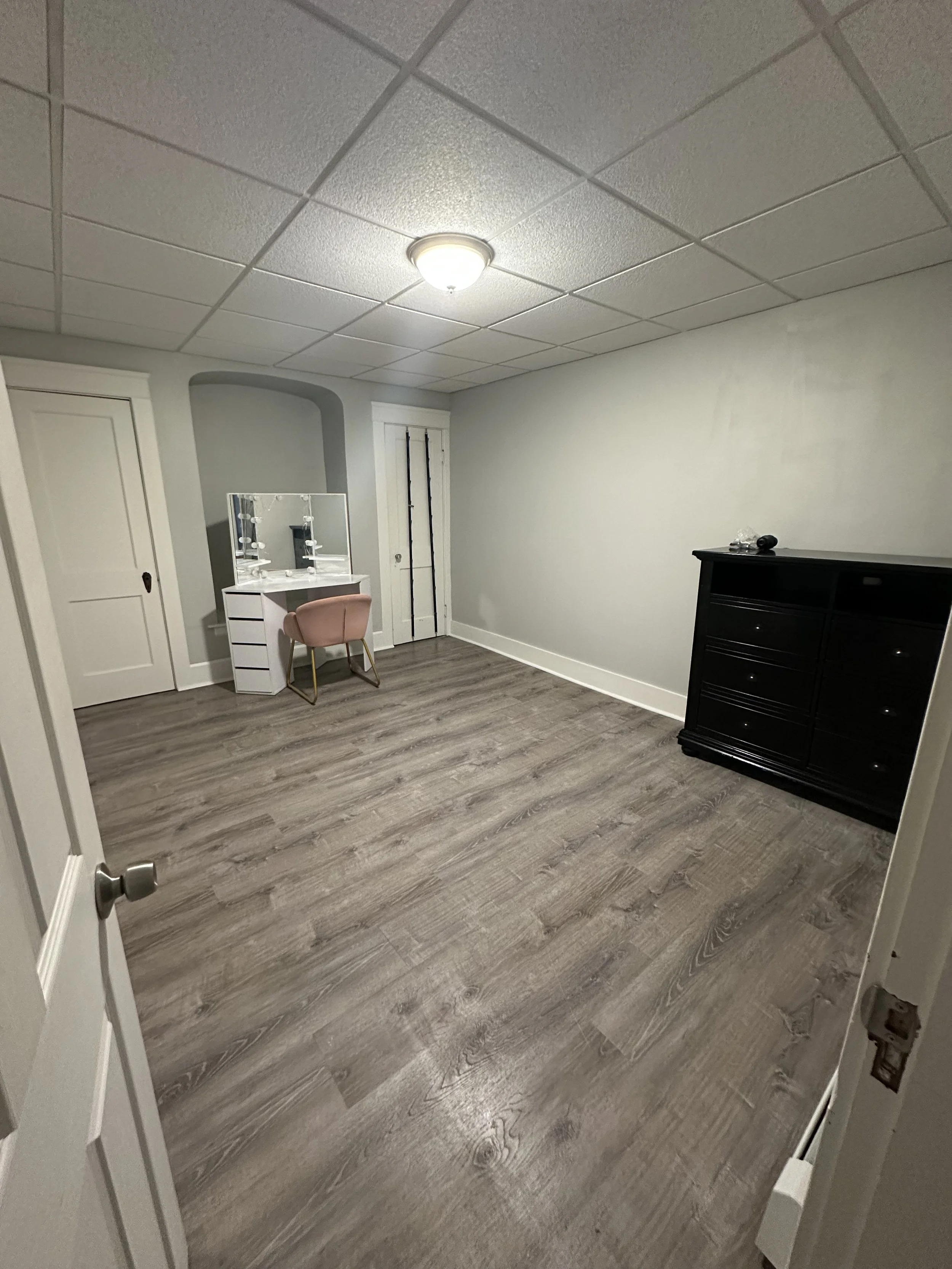 Empty room with wood laminate flooring, a white vanity with two mirrors and a pink chair, a black dresser, and a closed white door.