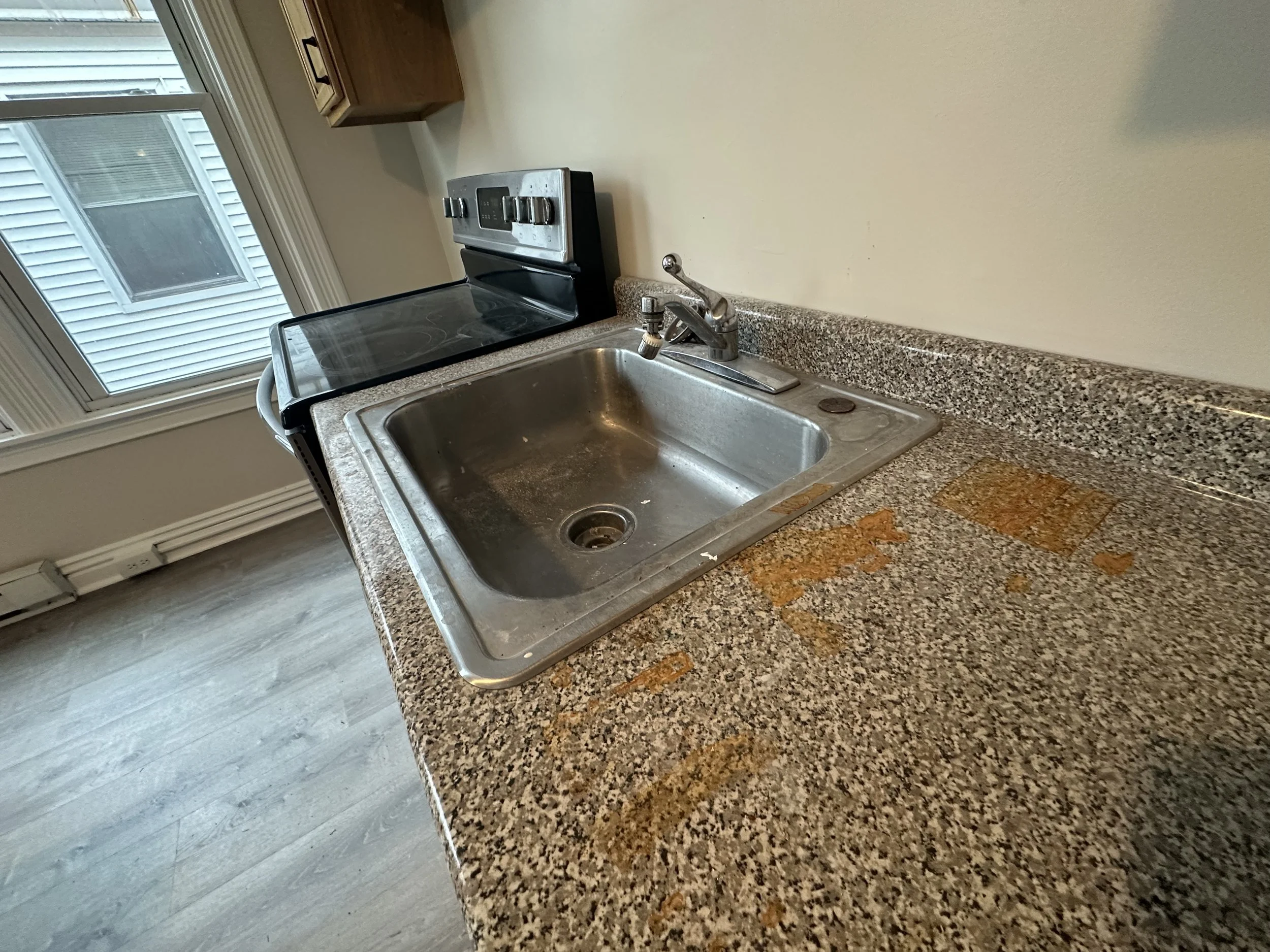 Dirty kitchen sink with rust stains on granite countertop, next to an electric stove, in a kitchen with a window overlooking an exterior wall.