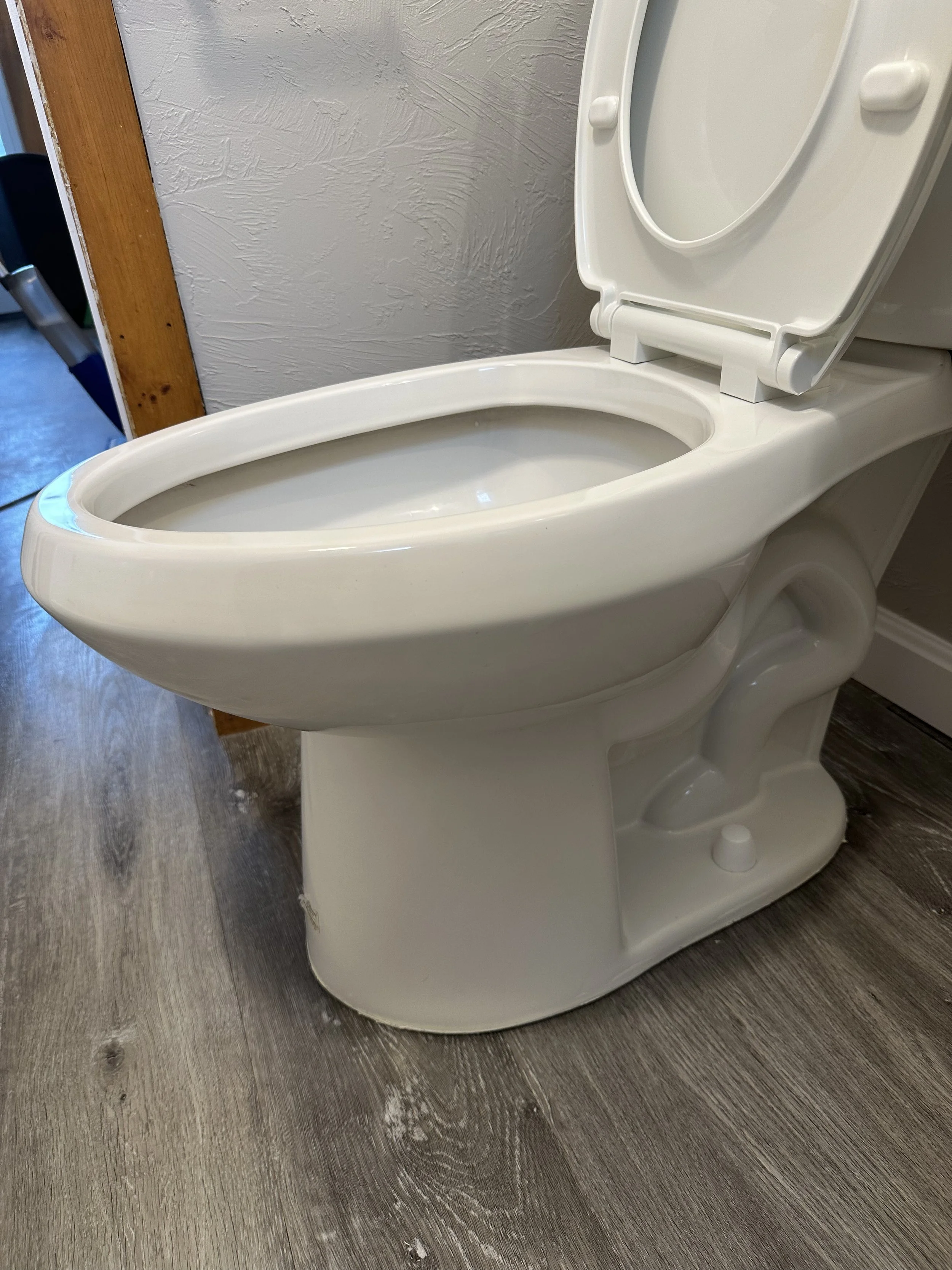A white ceramic toilet with the lid open, situated on a wooden floor next to a textured wall and a wooden door frame.