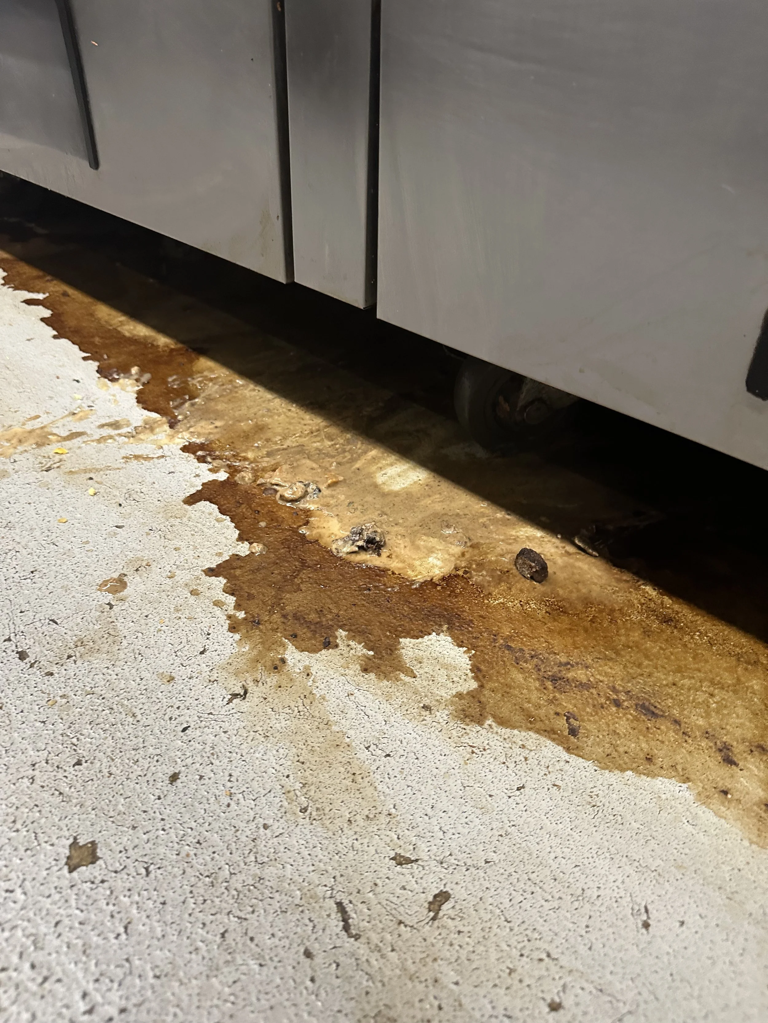 Oil spill and debris on concrete floor beneath a stainless steel appliance, possibly in a commercial kitchen.