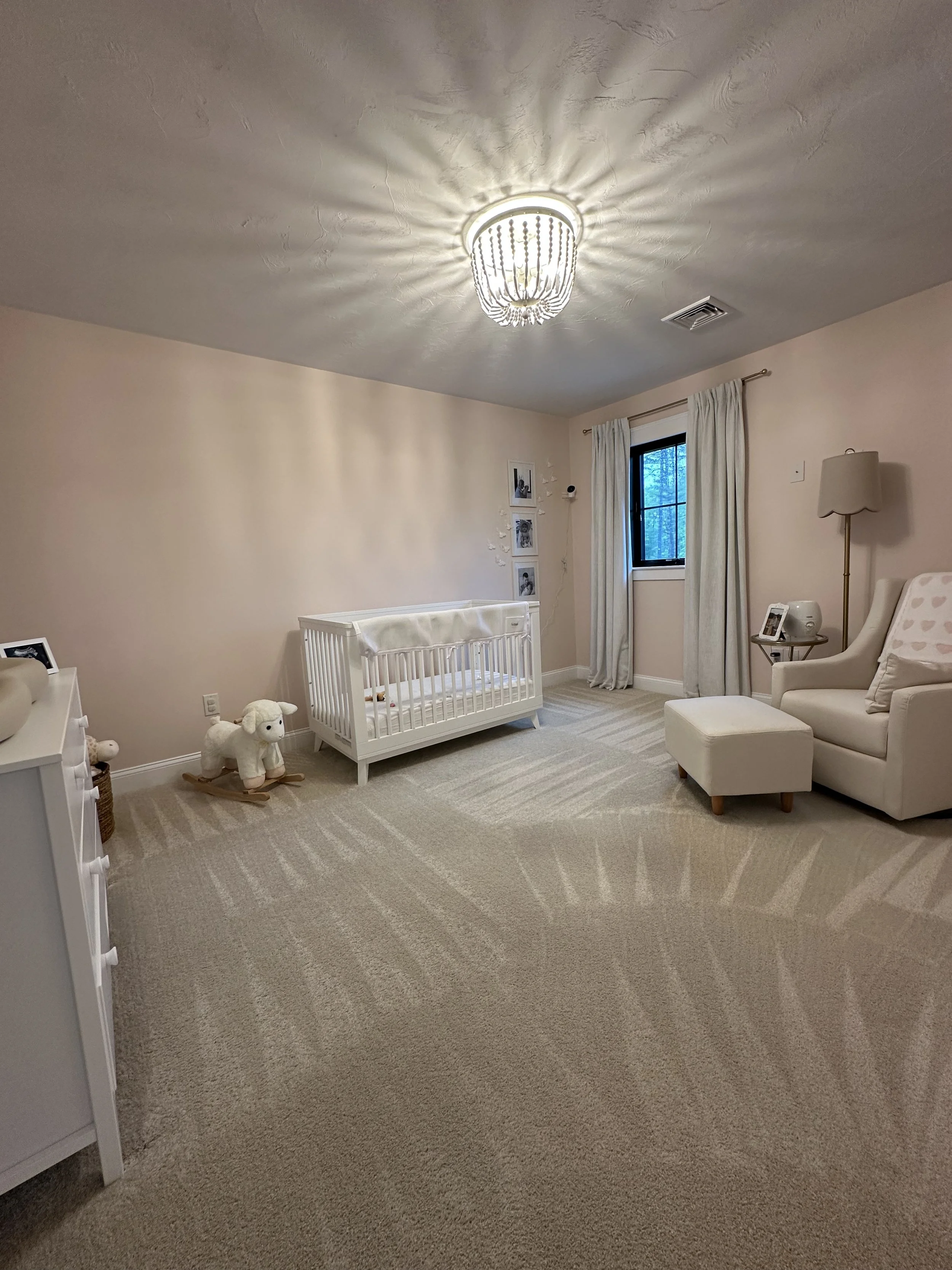 Nursery room with a white crib, soft toys, armchair, small ottoman, side table, window with curtains, and a chandelier.