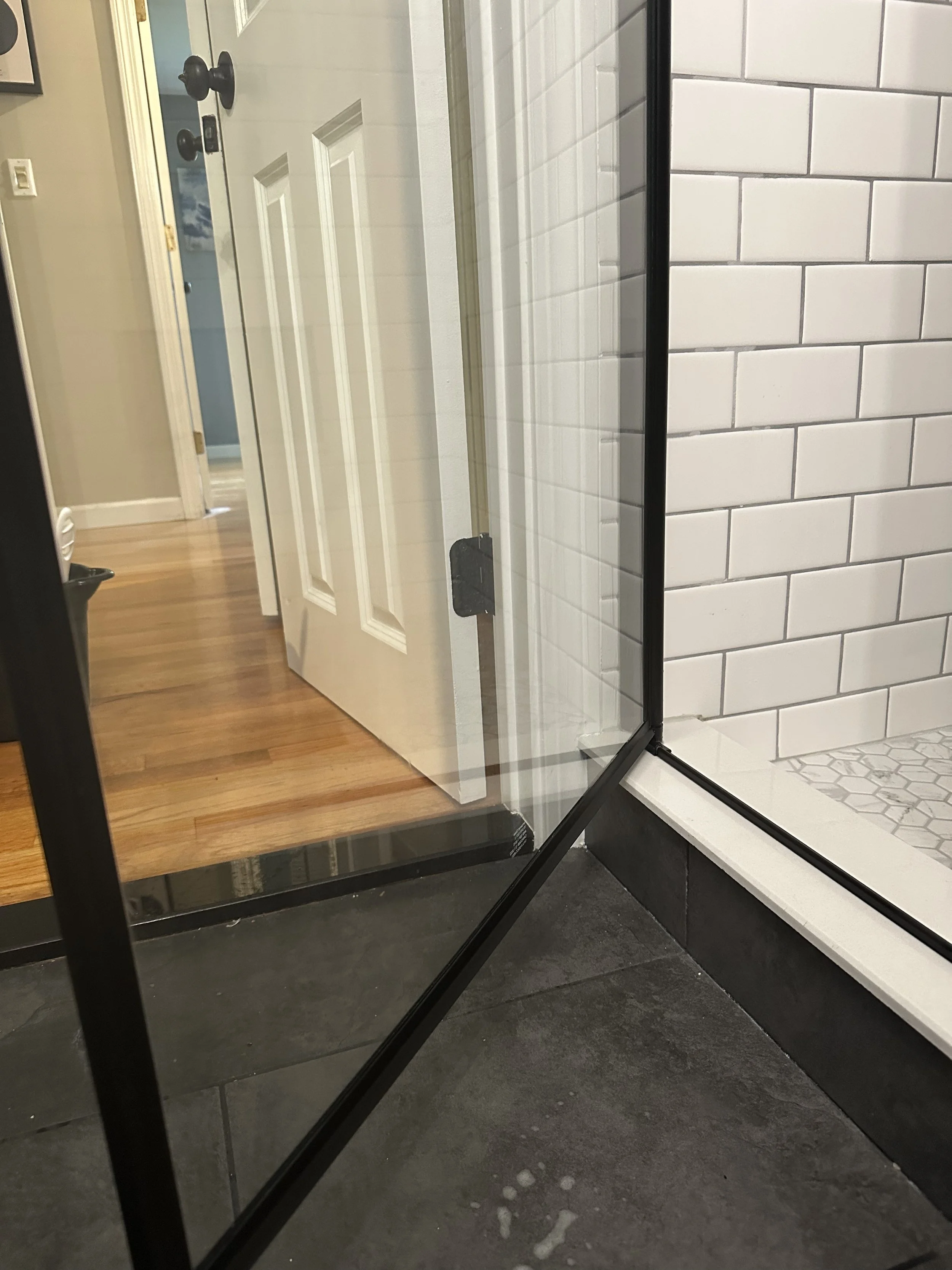 Close-up of a glass shower door with black metal frame next to a white subway tile shower wall. The reflection in the glass shows a wooden hallway, a white door with black knobs, and a beige wall.