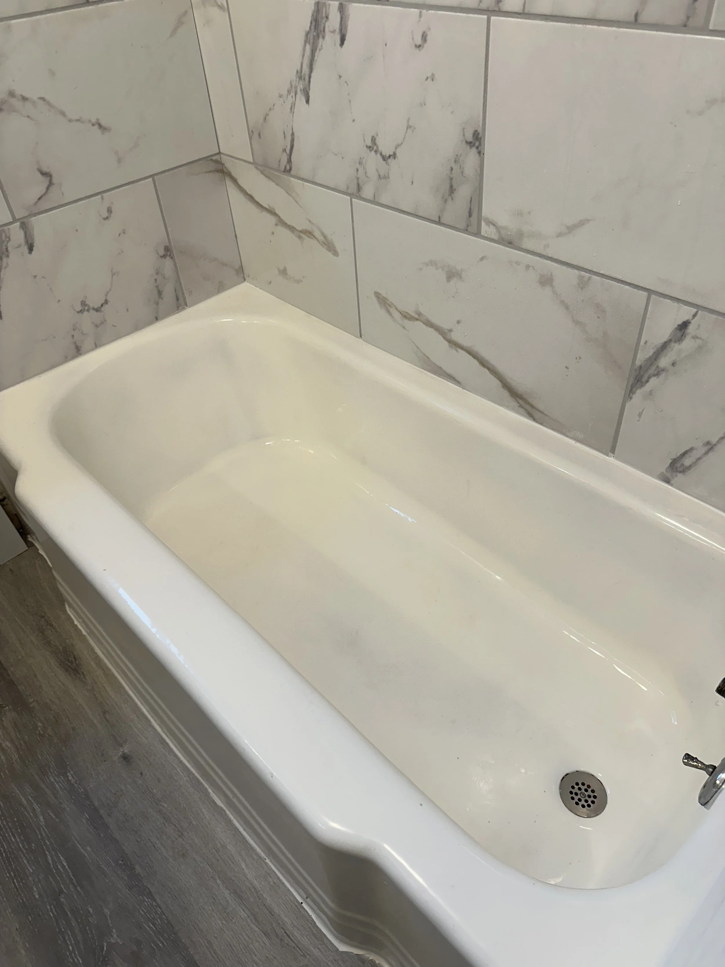 Empty white bathtub in a bathroom with marble tile walls and wooden flooring.