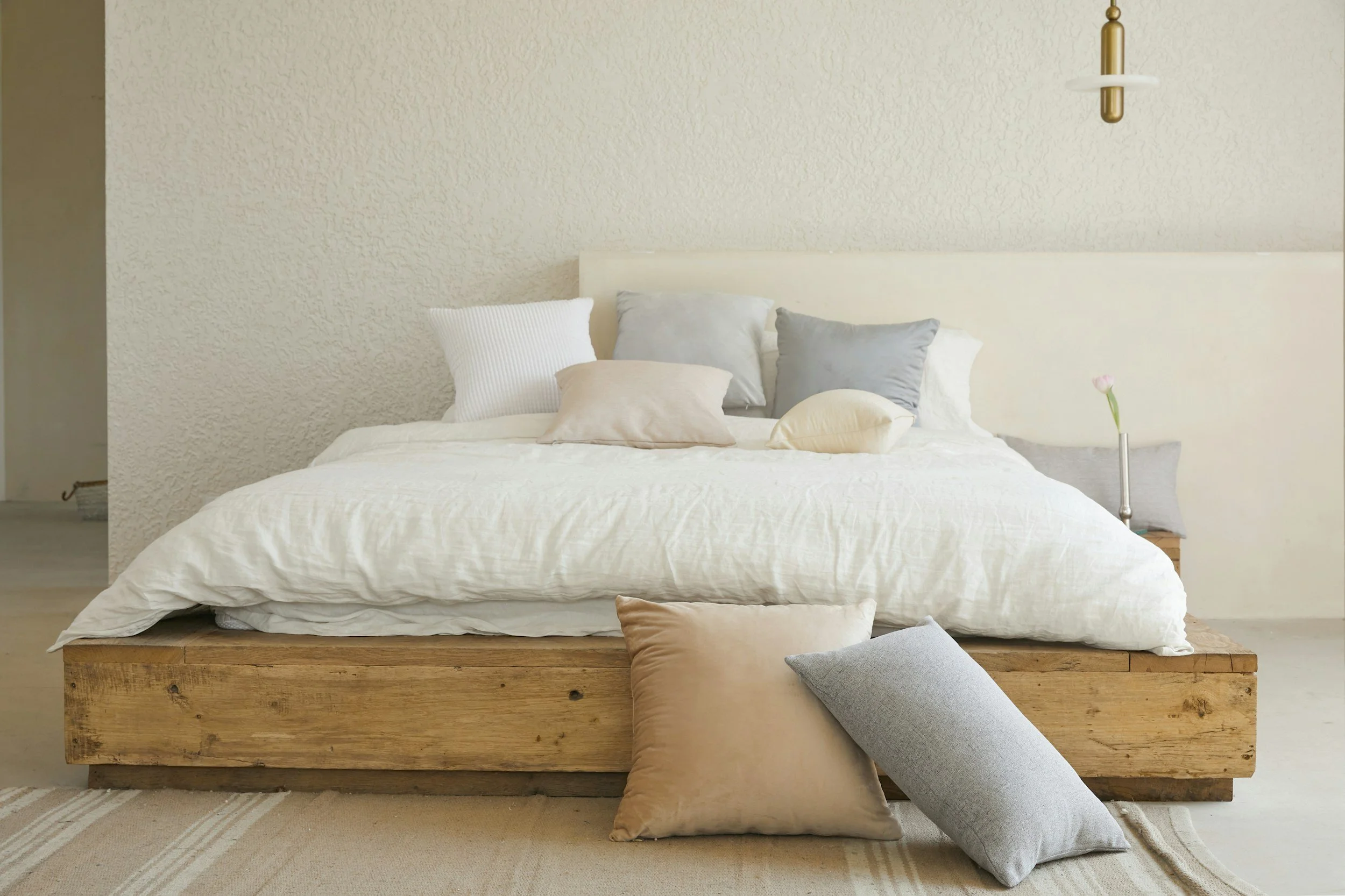 A bed with multiple beige, white, and gray pillows, a beige comforter, a wooden platform base, and a small bedside table with a flower in a glass vase in a minimalist bedroom.