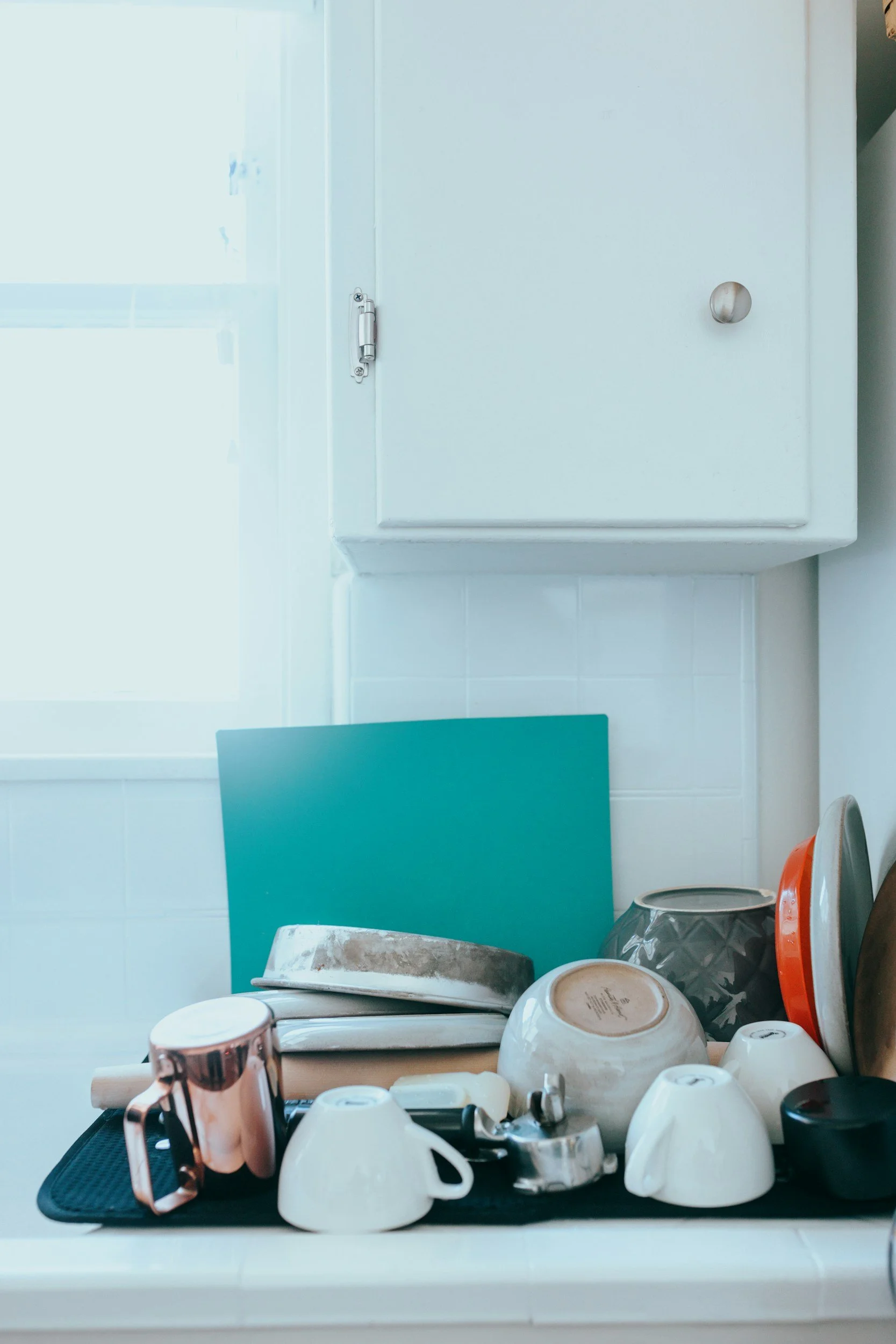 Kitchen countertop with various dishes and kitchenware, including bowls, cups, a mug, a piece of teal cutting board, and a metal pot, near a white cabinet and a window.