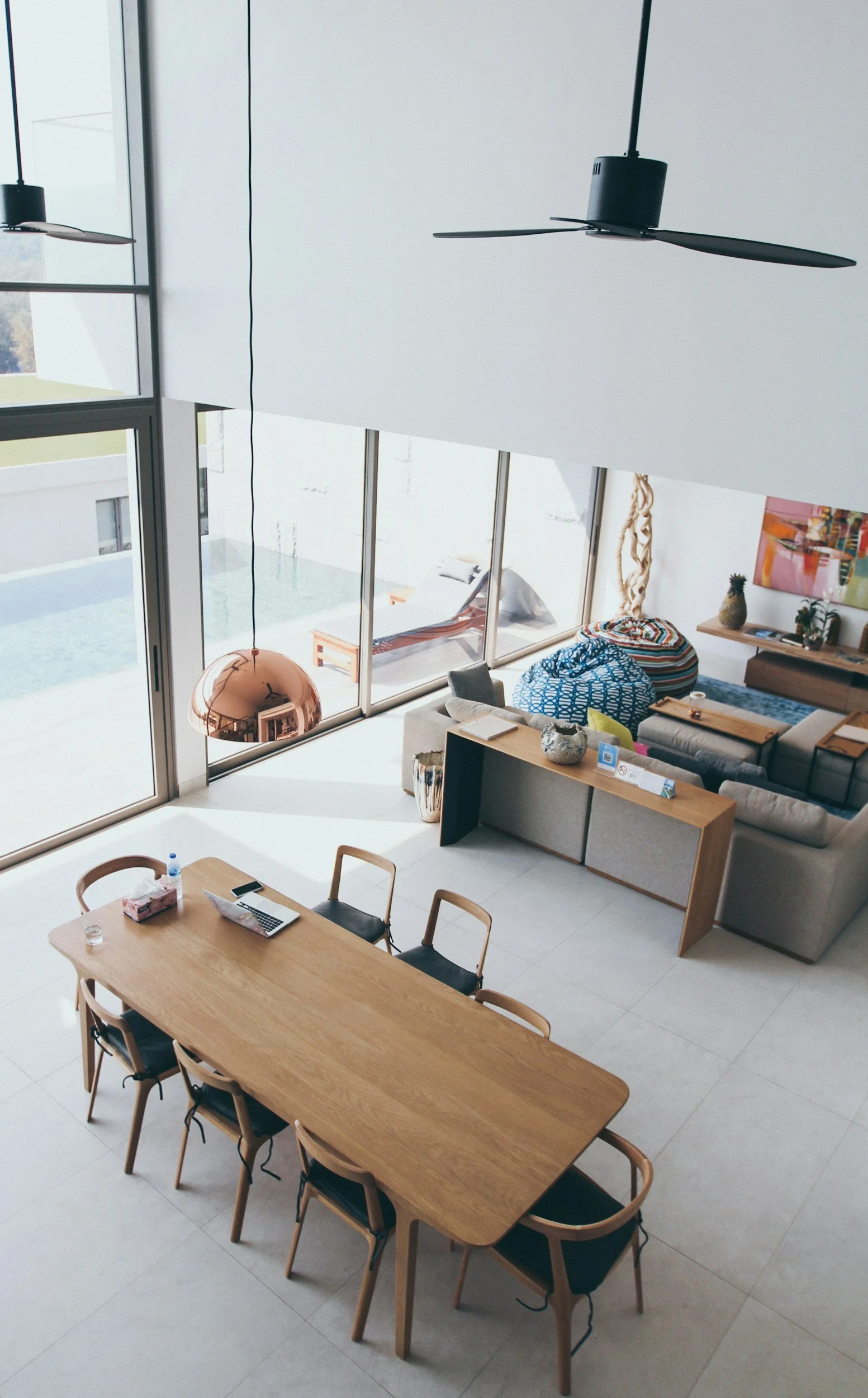 Interior of a modern, open-concept living and dining area with a wooden dining table, six chairs, a sofa, colorful cushions, a sideboard, and large floor-to-ceiling windows.