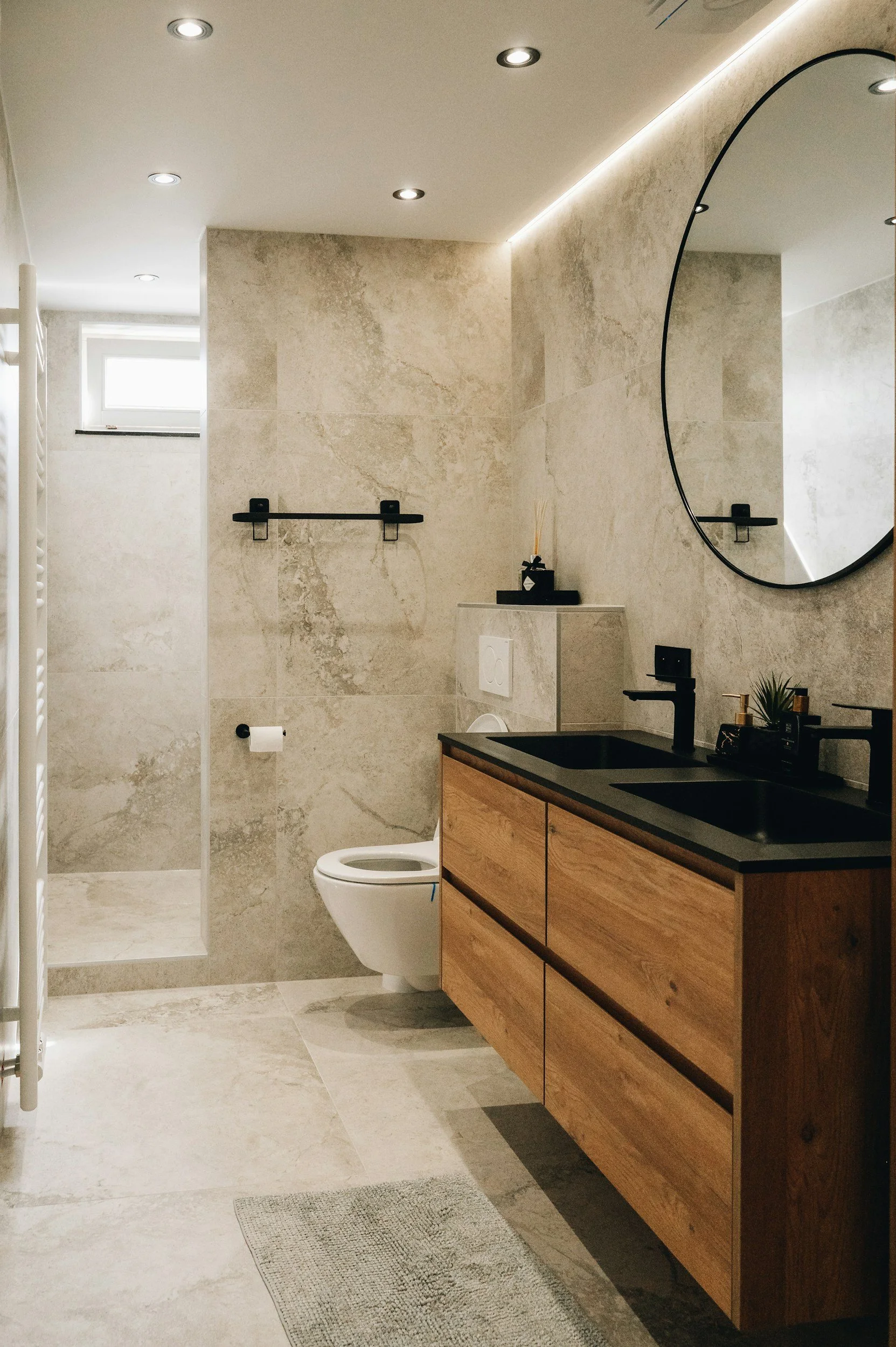Modern bathroom featuring a double sink vanity with black countertop, round mirror, wall-mounted toilet, frosted window, black towel rack, small shelf with decor, and a shower area in the background with natural stone tiles.