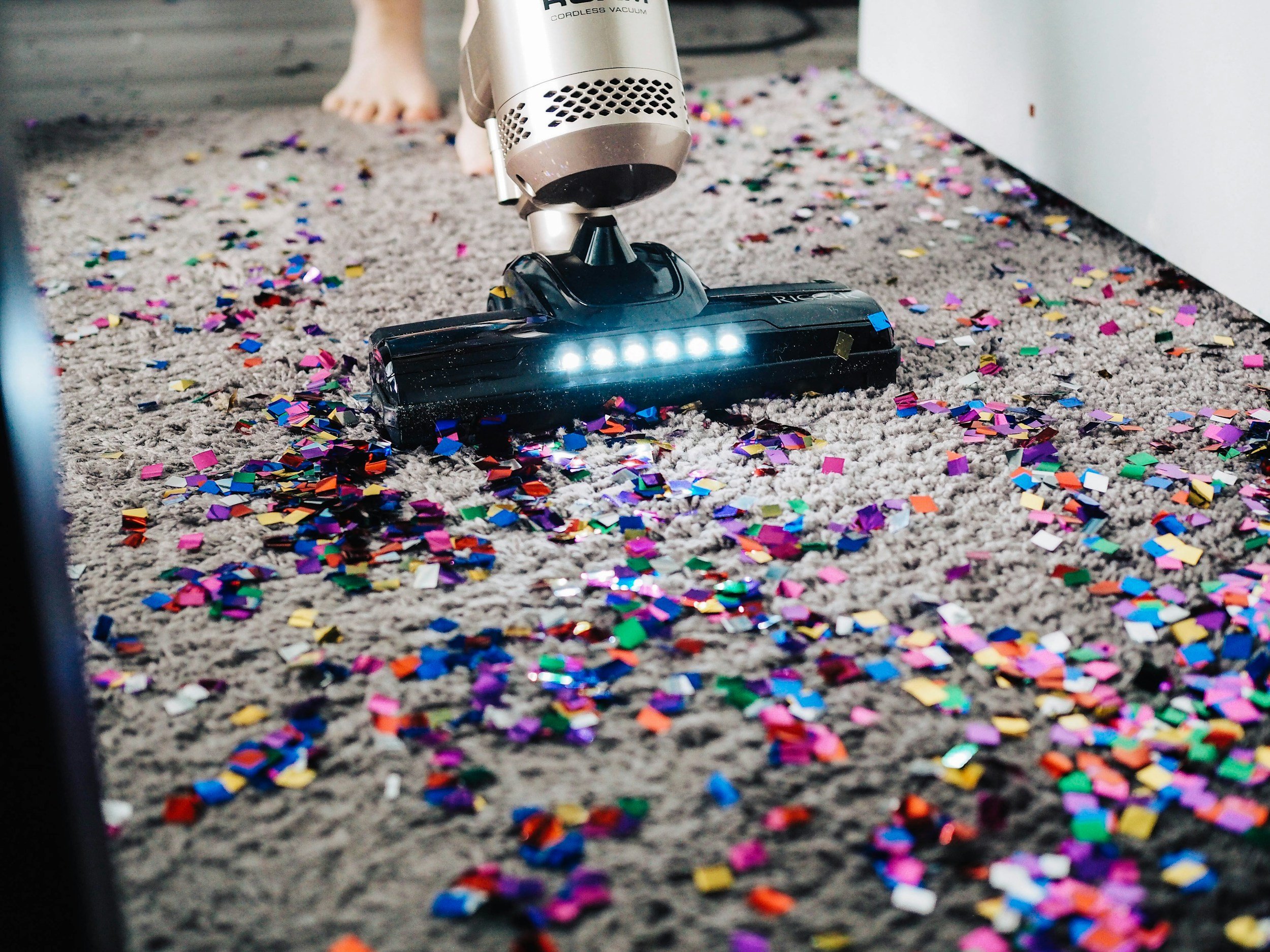 A vacuum cleaner is cleaning up colorful confetti on a gray carpet.