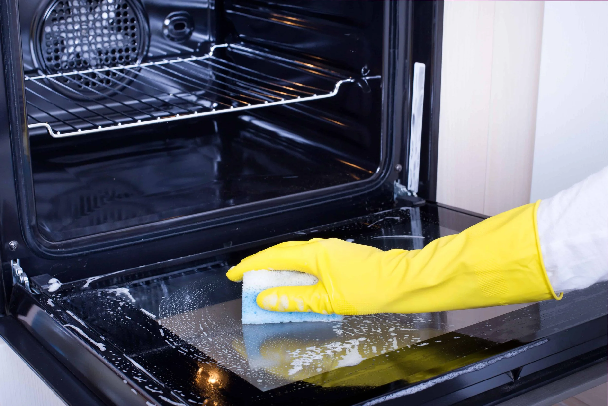 Person wearing yellow rubber gloves cleaning glass oven door with a sponge and soap suds.